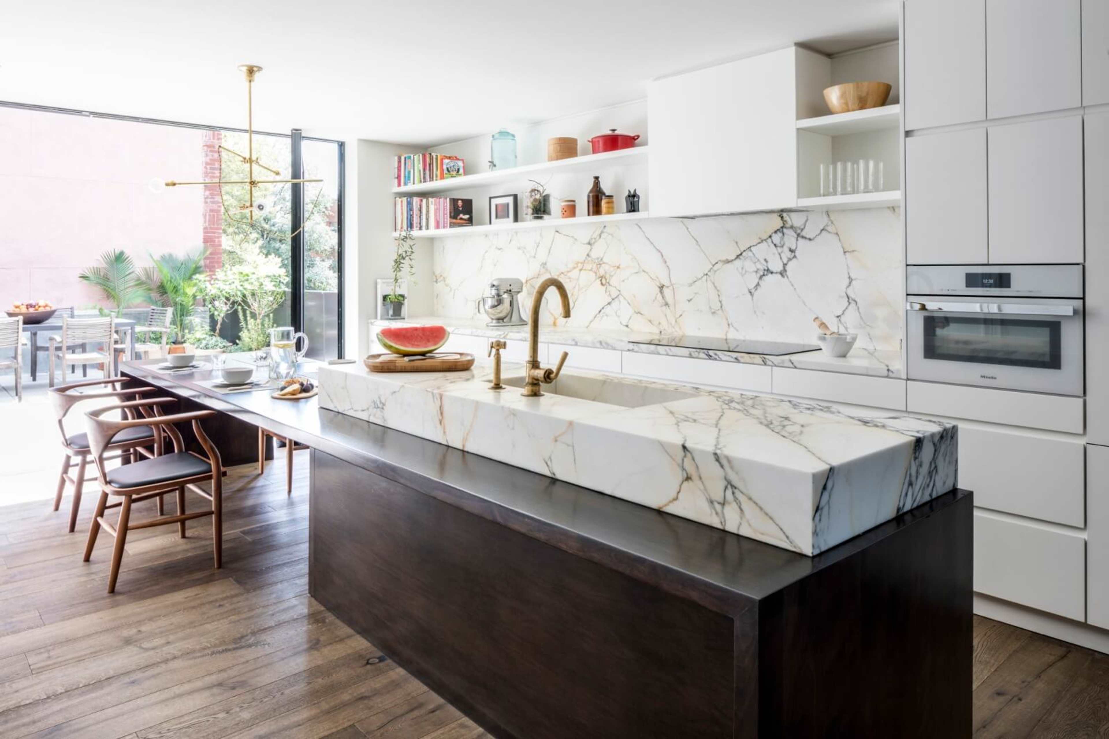 A modern kitchen with a large marble island and a wooden dining table, featuring an outdoor patio visible through large sliding glass doors.