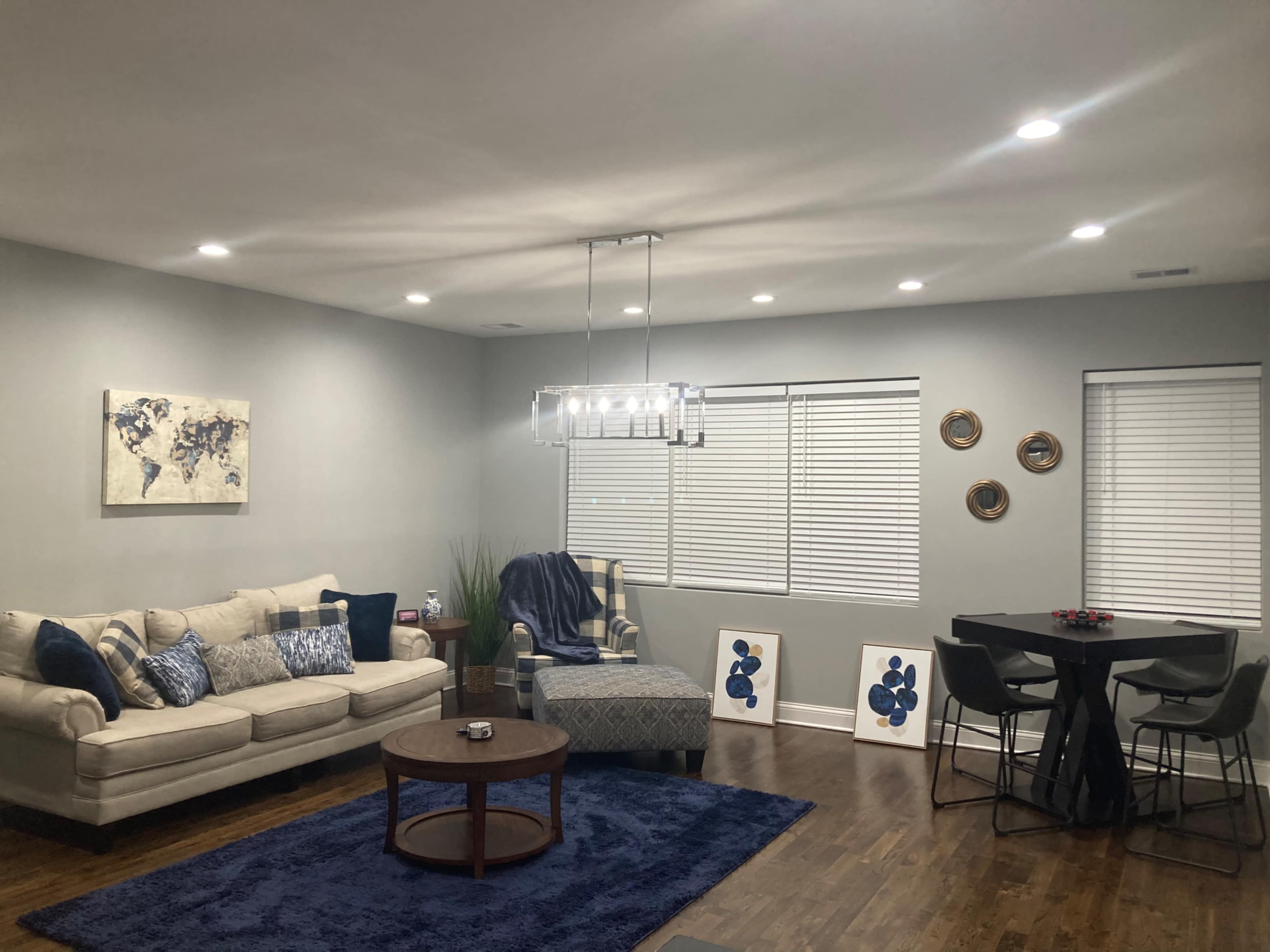 A modern living room with a beige sectional couch, a round wooden coffee table, a dining area with a black table and chairs, and decorative artwork on the walls.
