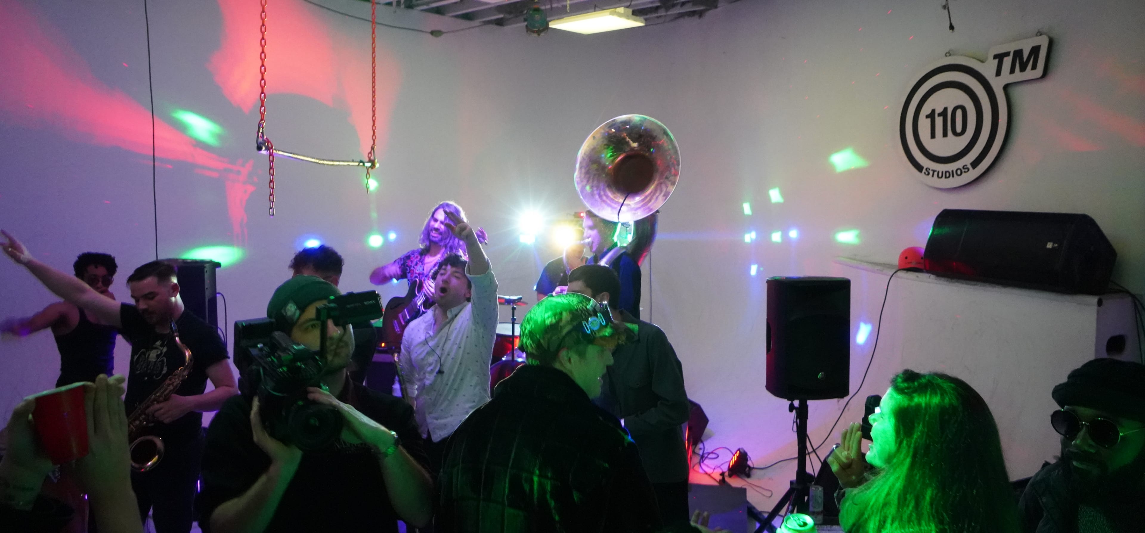 A lively gathering takes place in a studio with colorful lights illuminating people dancing and playing musical instruments.