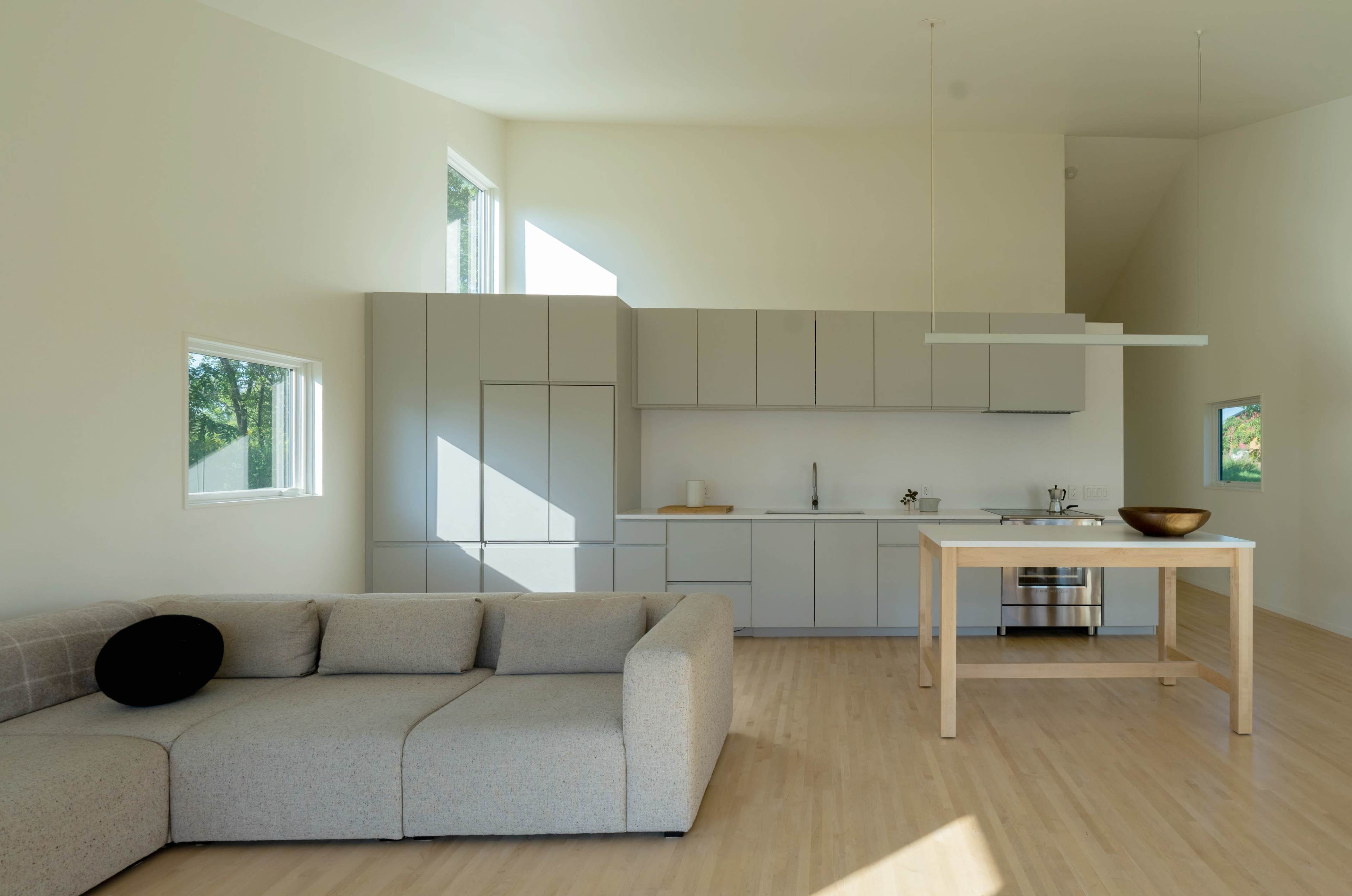 The image shows a modern, open-concept kitchen and living area featuring a light-colored sofa and minimalist cabinetry.