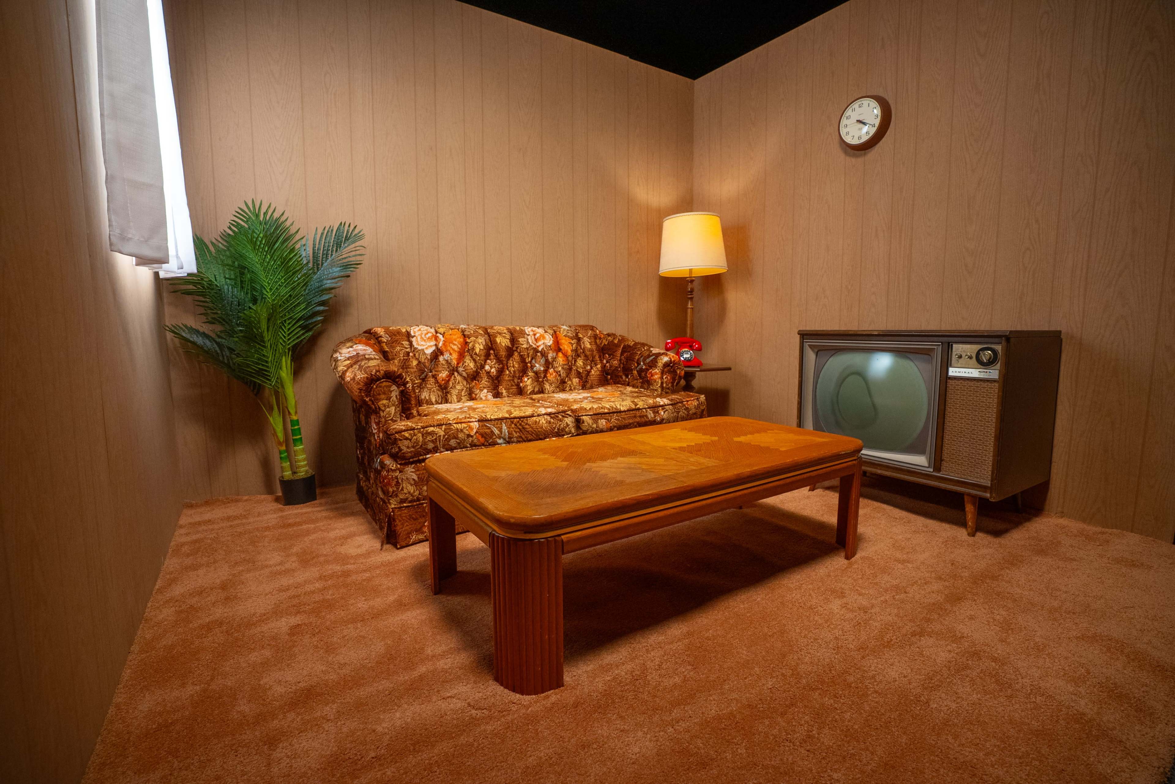 The image shows a retro-styled living room with a patterned couch, a wooden coffee table, a vintage television, a lamp, and a potted plant.