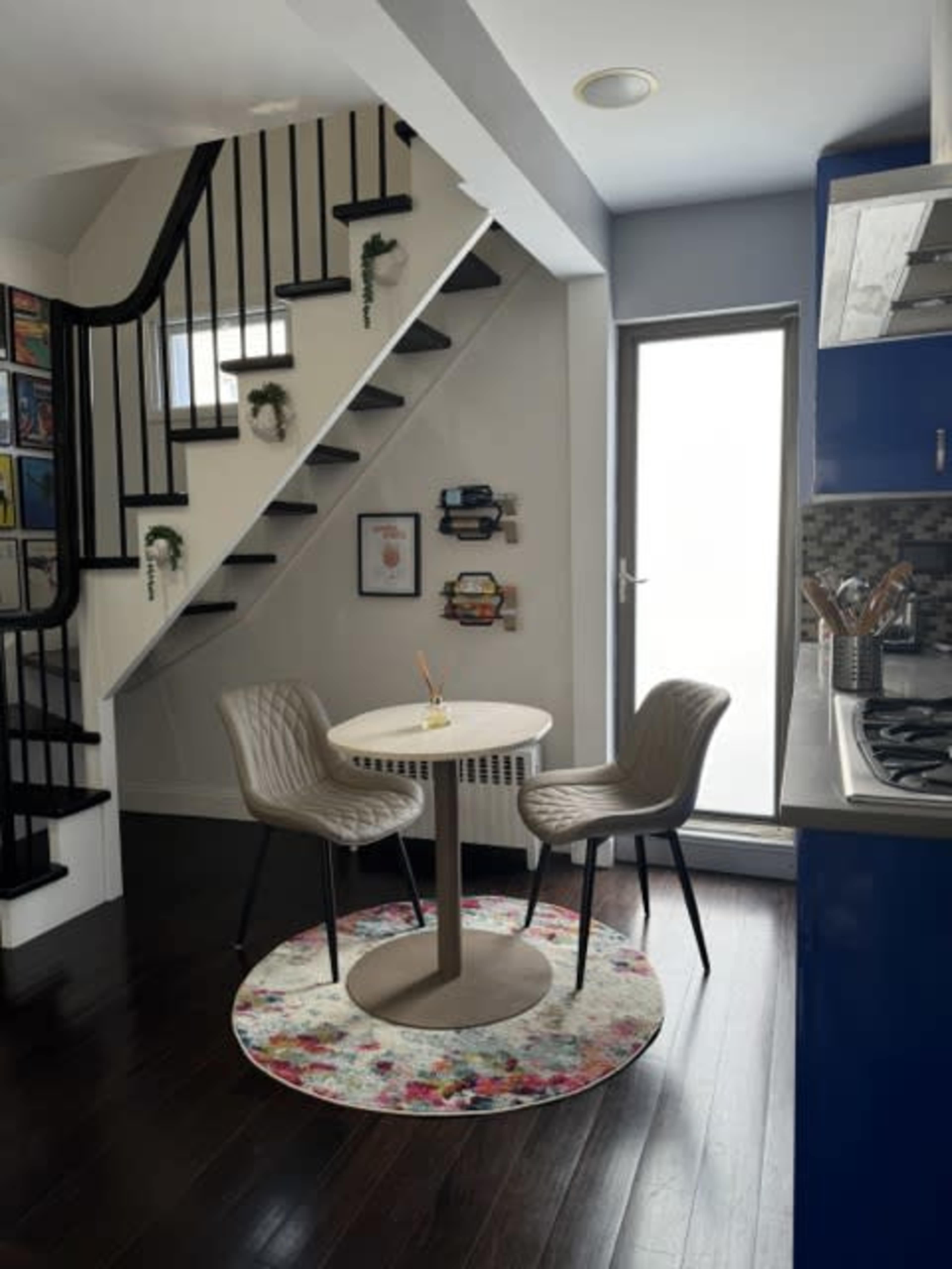 A small dining area features a circular table with two chairs, situated next to a staircase and framed by a colorful rug.