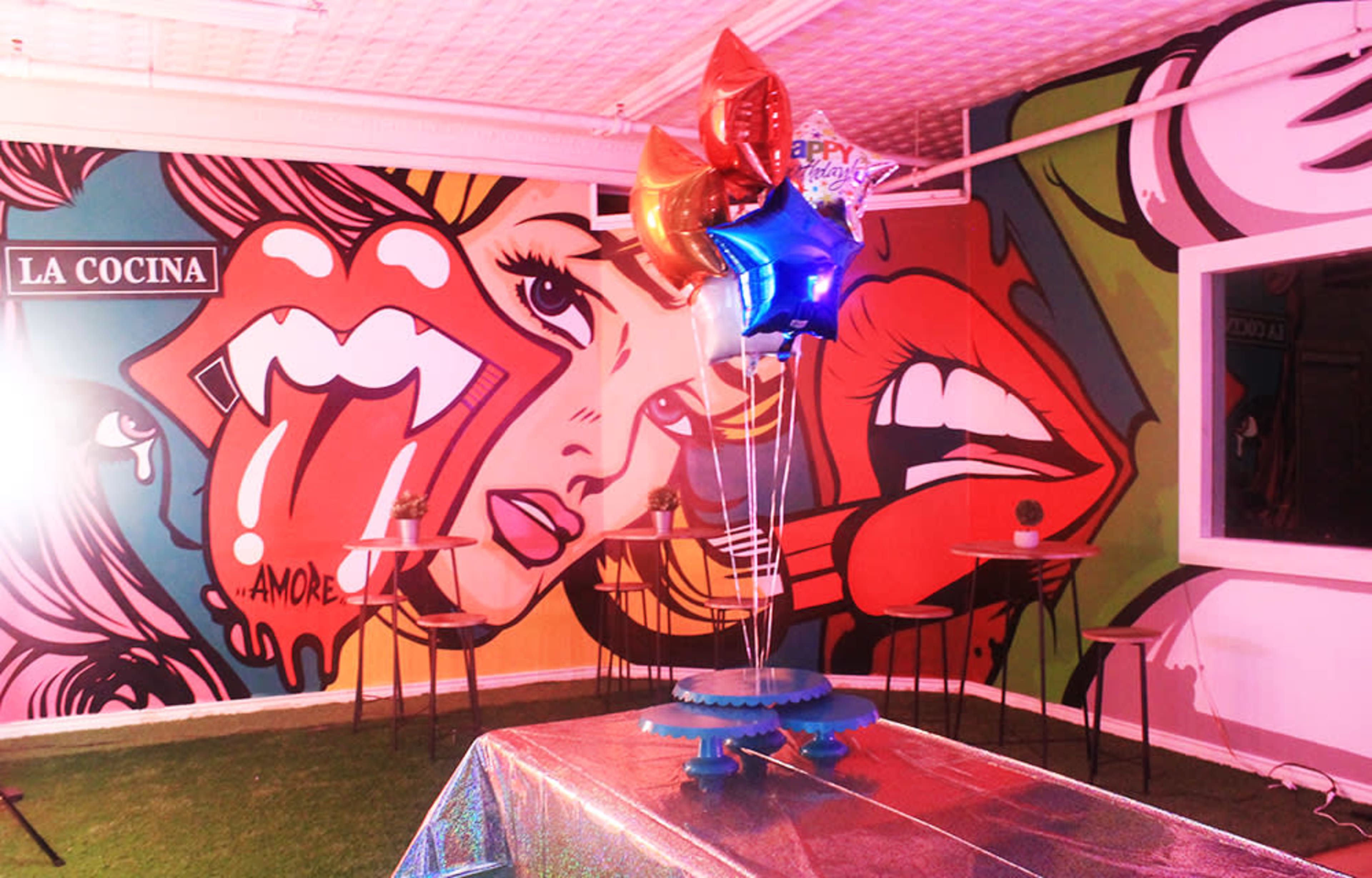 A colorful mural featuring stylized lips and a tongue adorns the wall of a room with a table decorated for a celebration, complete with balloons and a shiny tablecloth.