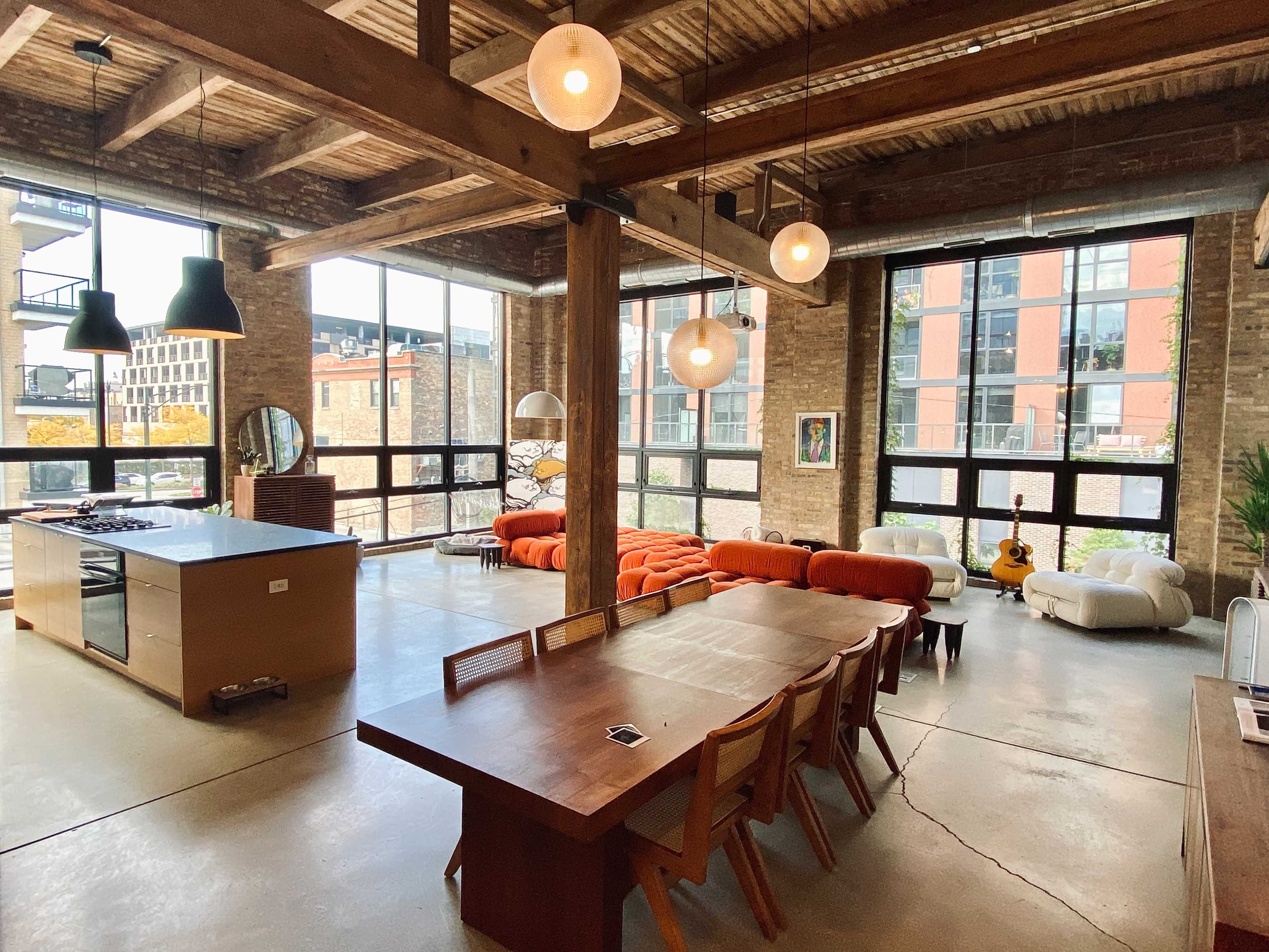 A modern loft features an open layout with a dining table, kitchen island, and various seating areas, all illuminated by pendant lights and large windows.