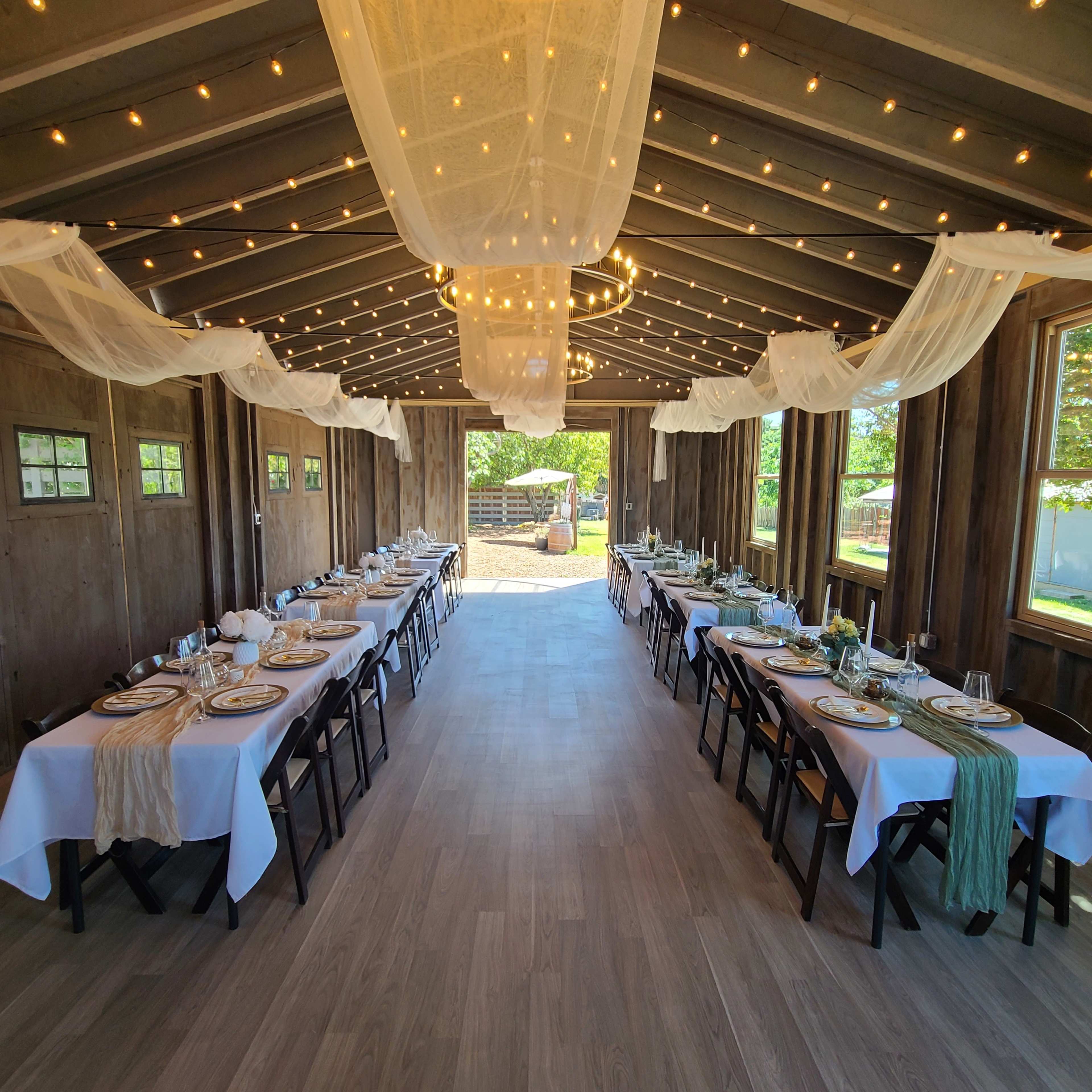 A spacious indoor dining area is set up with two long tables adorned with tableware and greenery, surrounded by strings of lights and draped fabric, leading to an outdoor view.