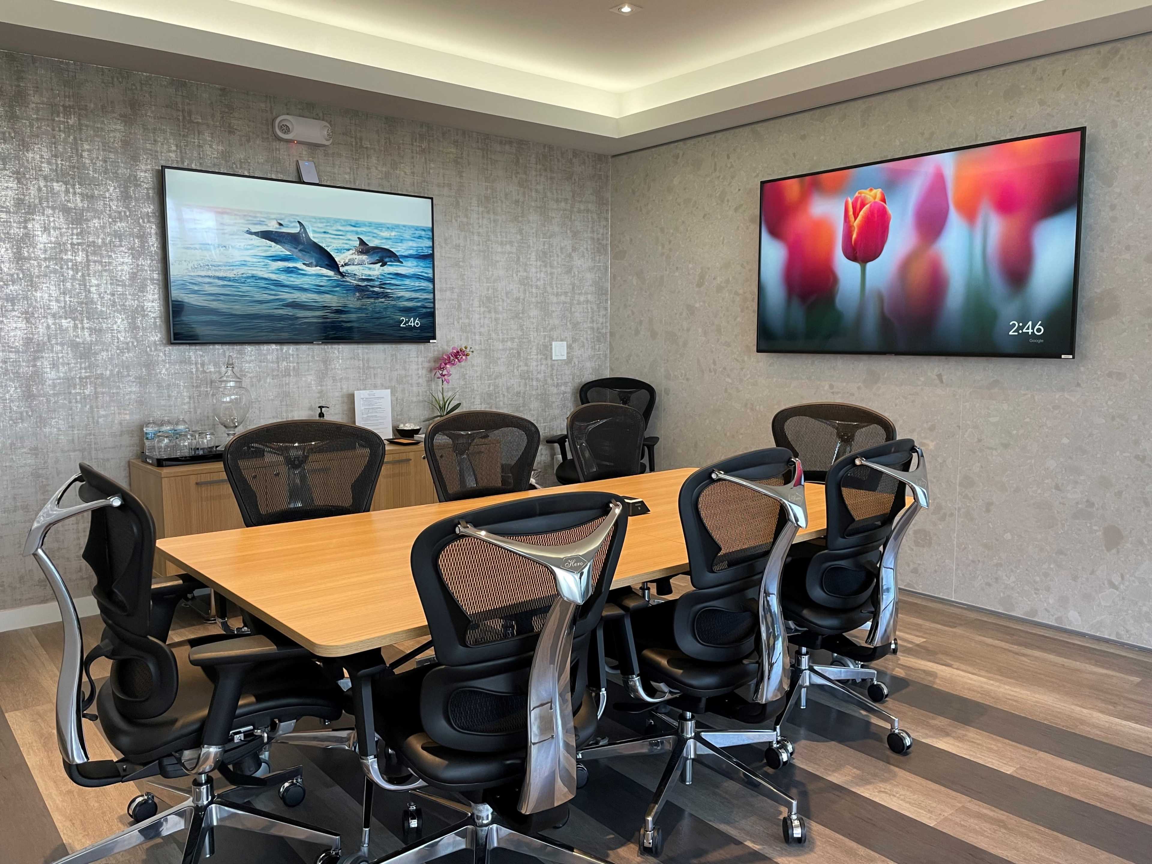 A modern conference room features a large wooden table surrounded by ergonomic chairs, with two screens displaying images on the walls.