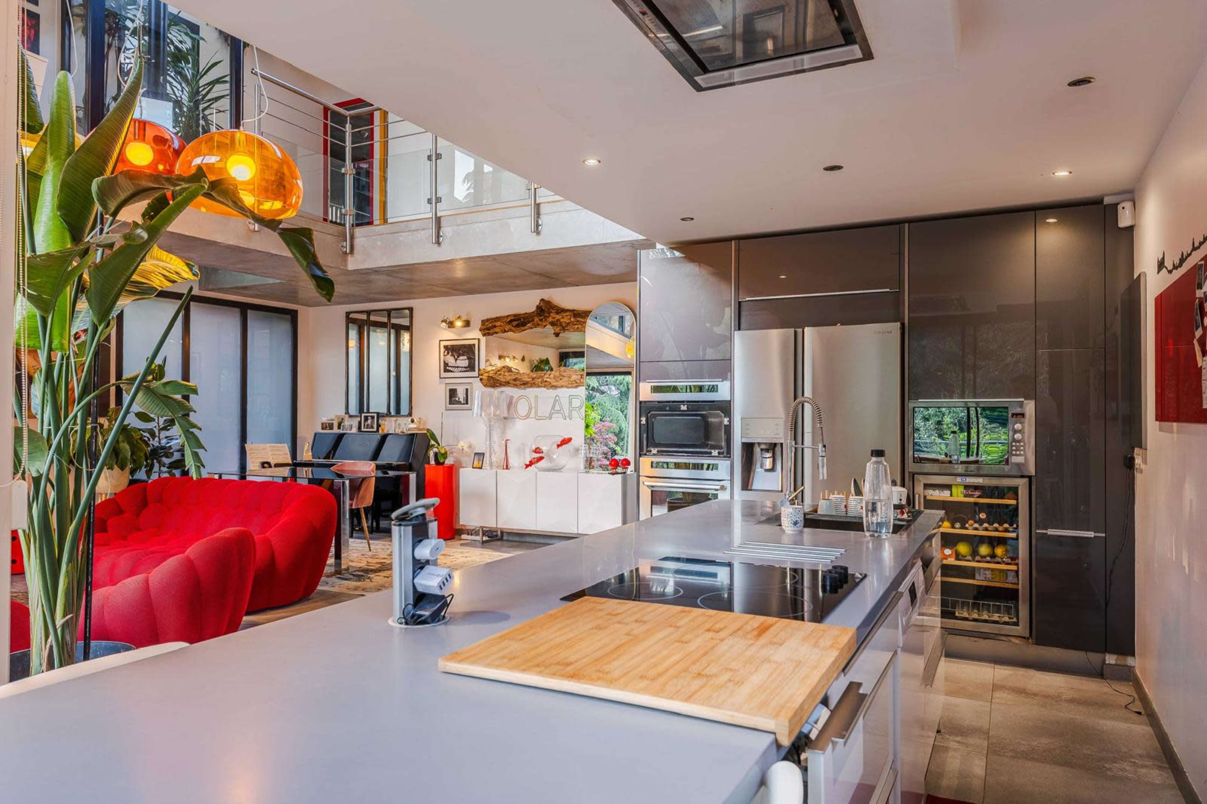 The image shows a modern kitchen with a sleek countertop, stainless steel appliances, and an open layout that connects to a living area featuring red seating.