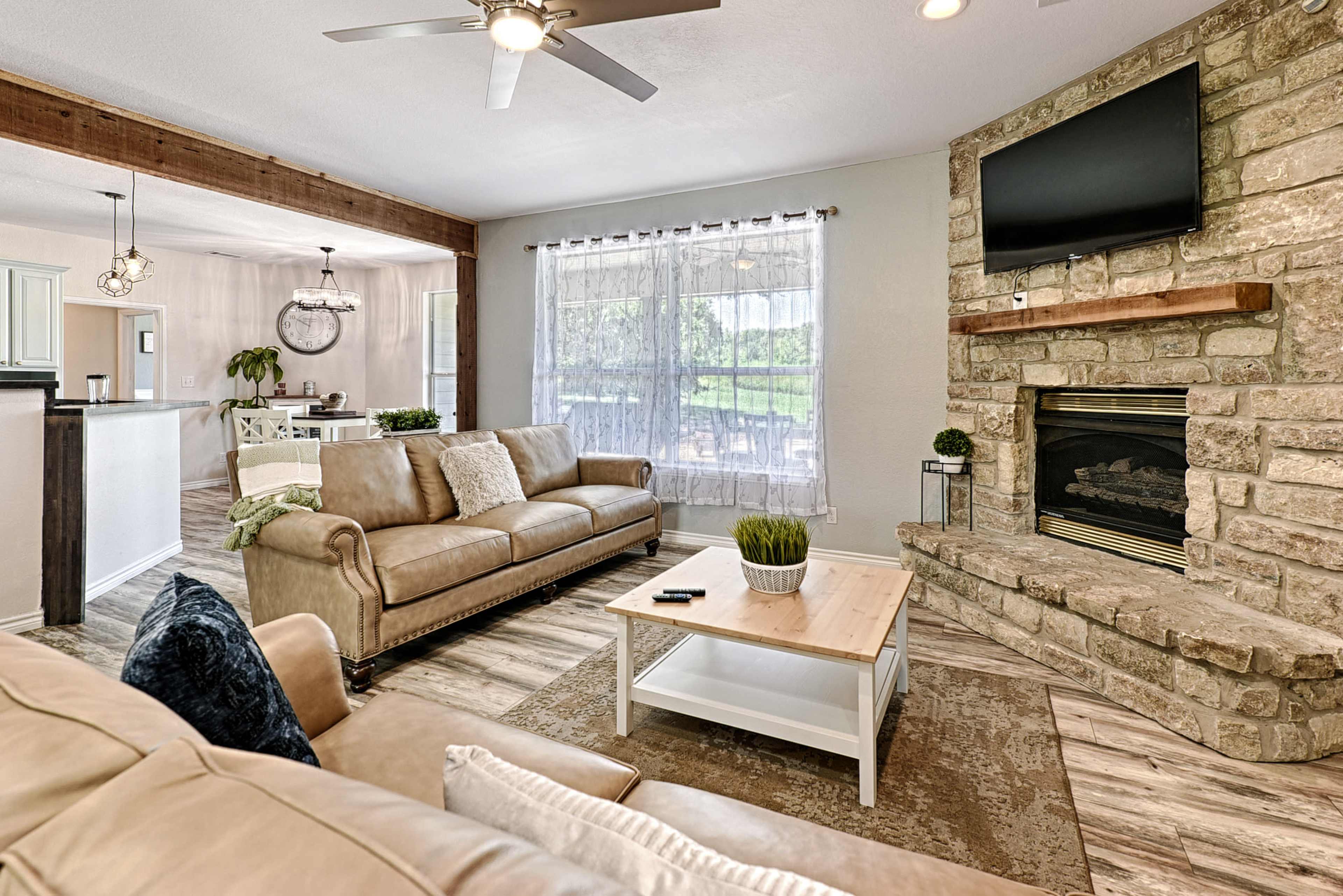 The image shows a modern living room with a stone fireplace, two beige sofas, a coffee table, and large windows that let in natural light.