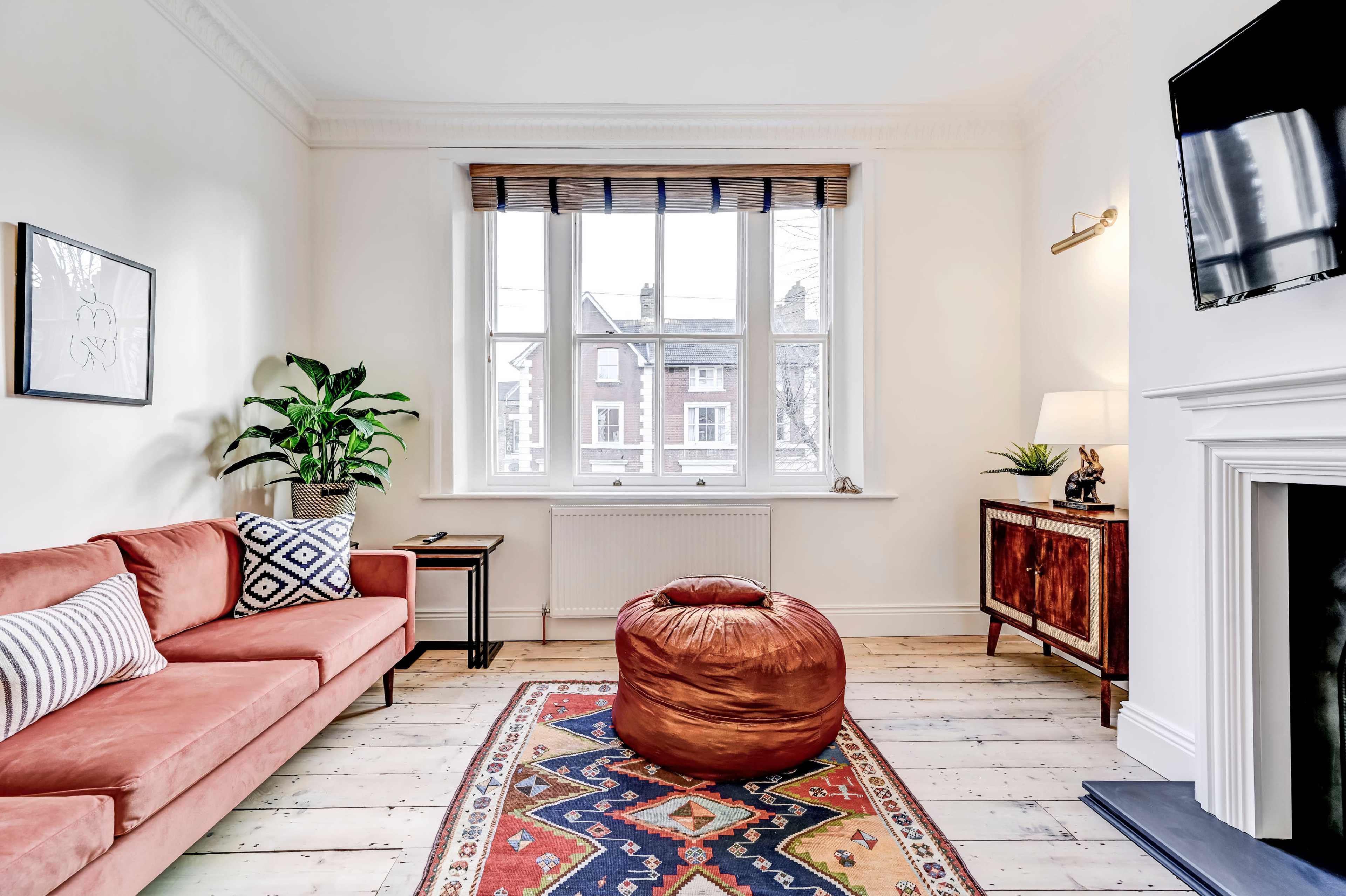 A living room features a pink sofa, a decorative rug, a large window with wooden blinds, a potted plant, and a cozy pouf.
