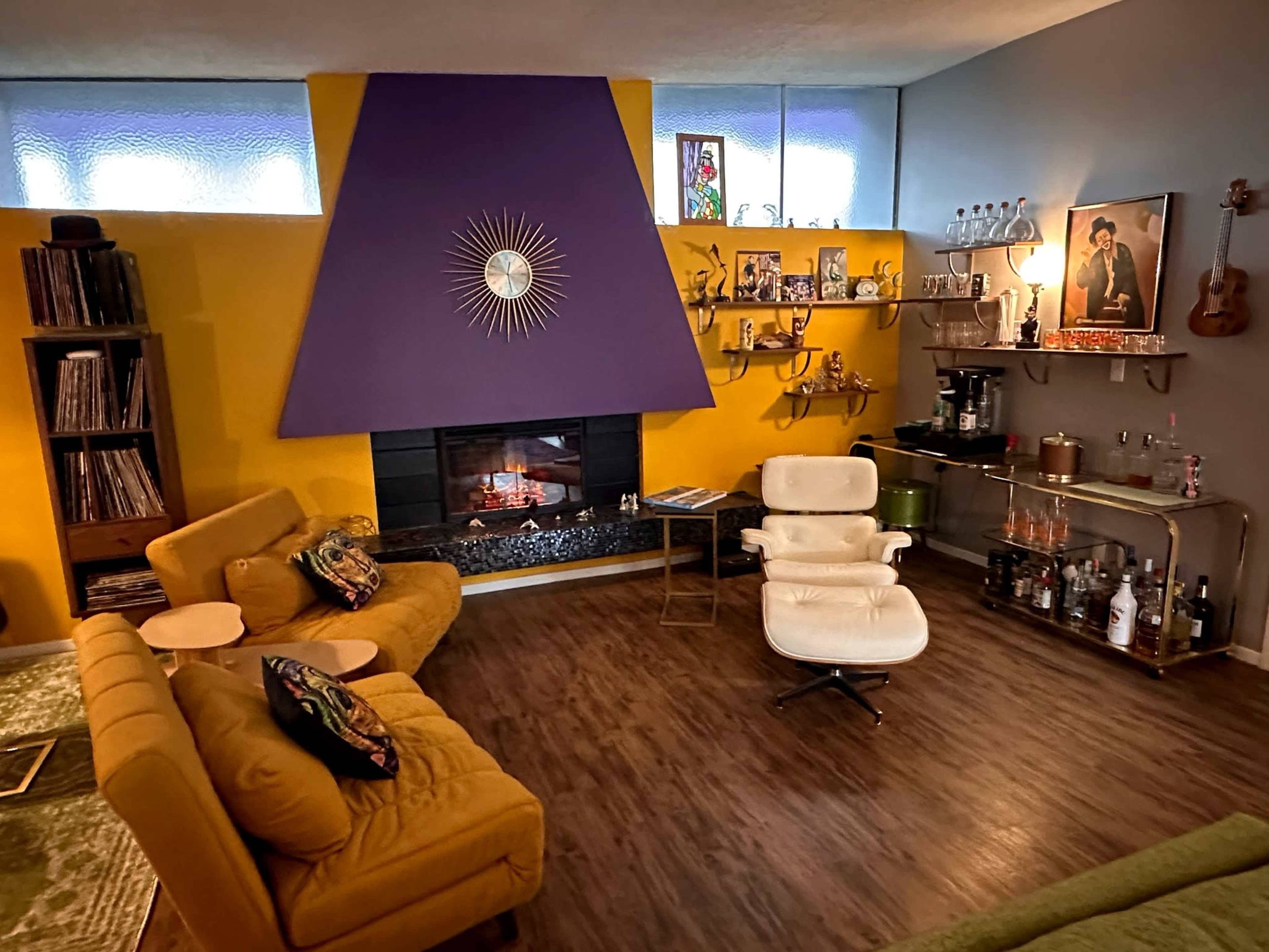 A cozy living room with a purple accent wall, a fireplace, yellow walls, and various furniture pieces including a white lounge chair and yellow sofas.