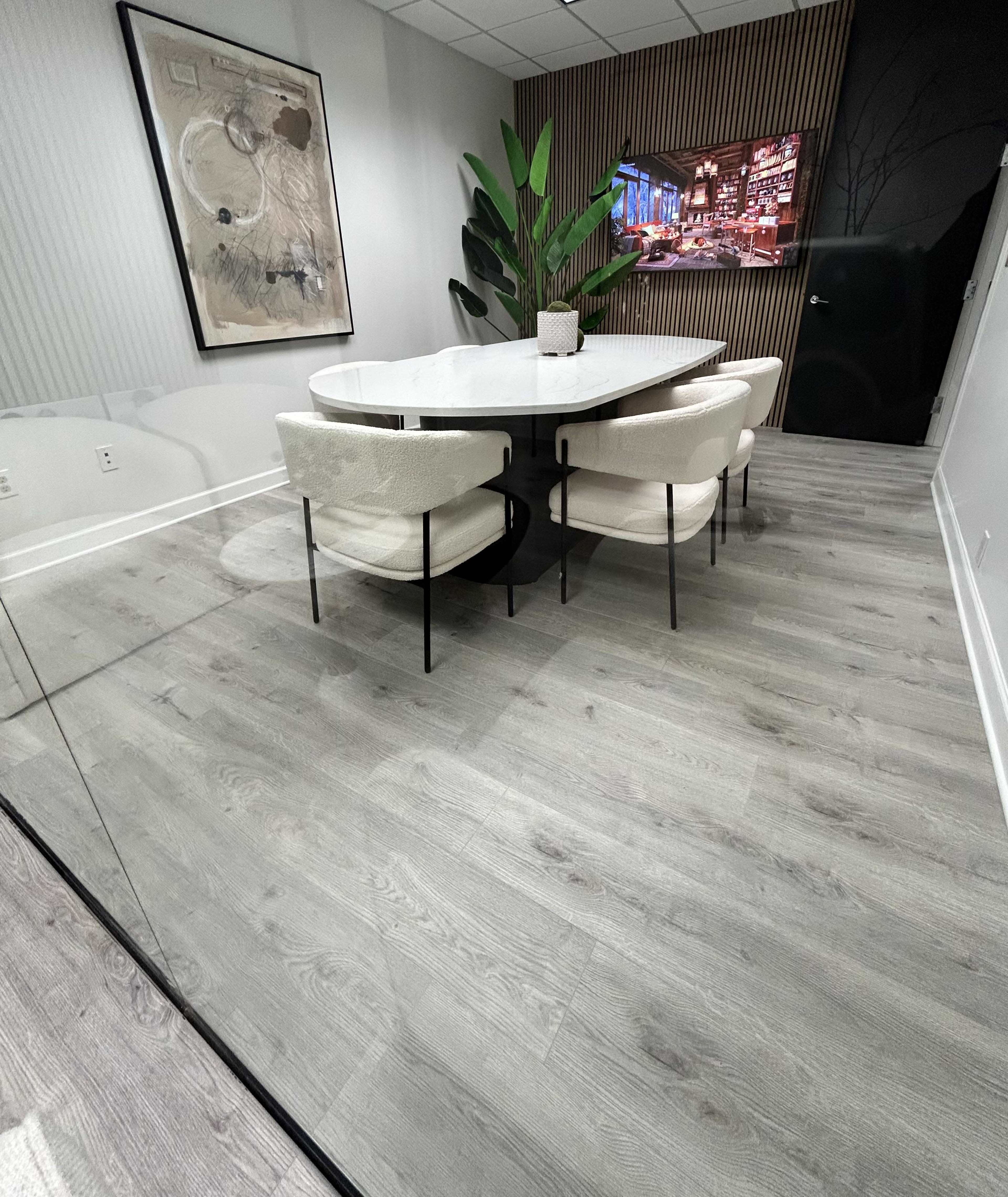 A modern conference room features a round table surrounded by four upholstered chairs, a large plant, and a wall-mounted screen displaying a city skyline.