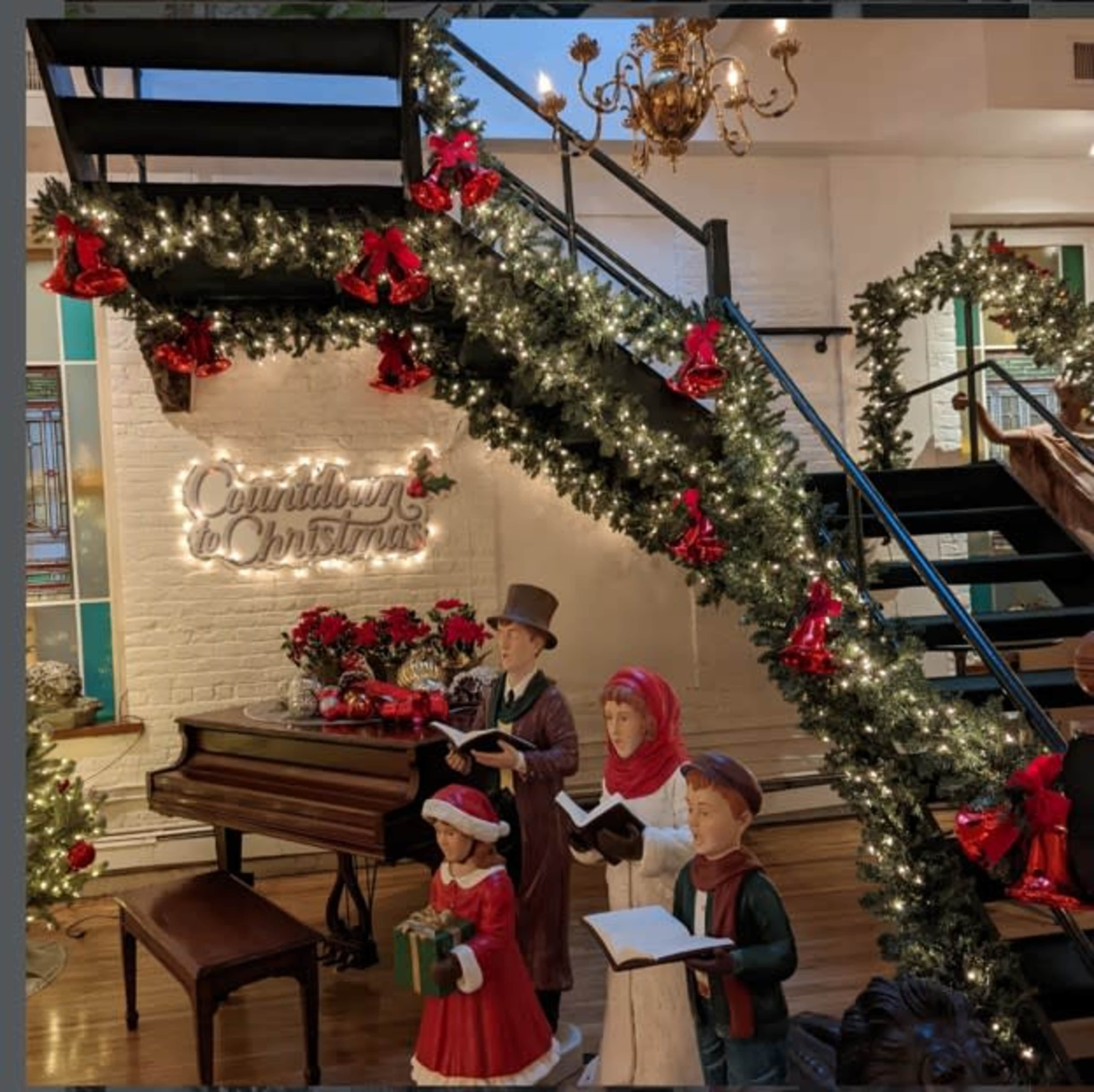 A decorated staircase features garlands, red bows, and a sign that reads "Countdown to Christmas," alongside festive figures and a piano.