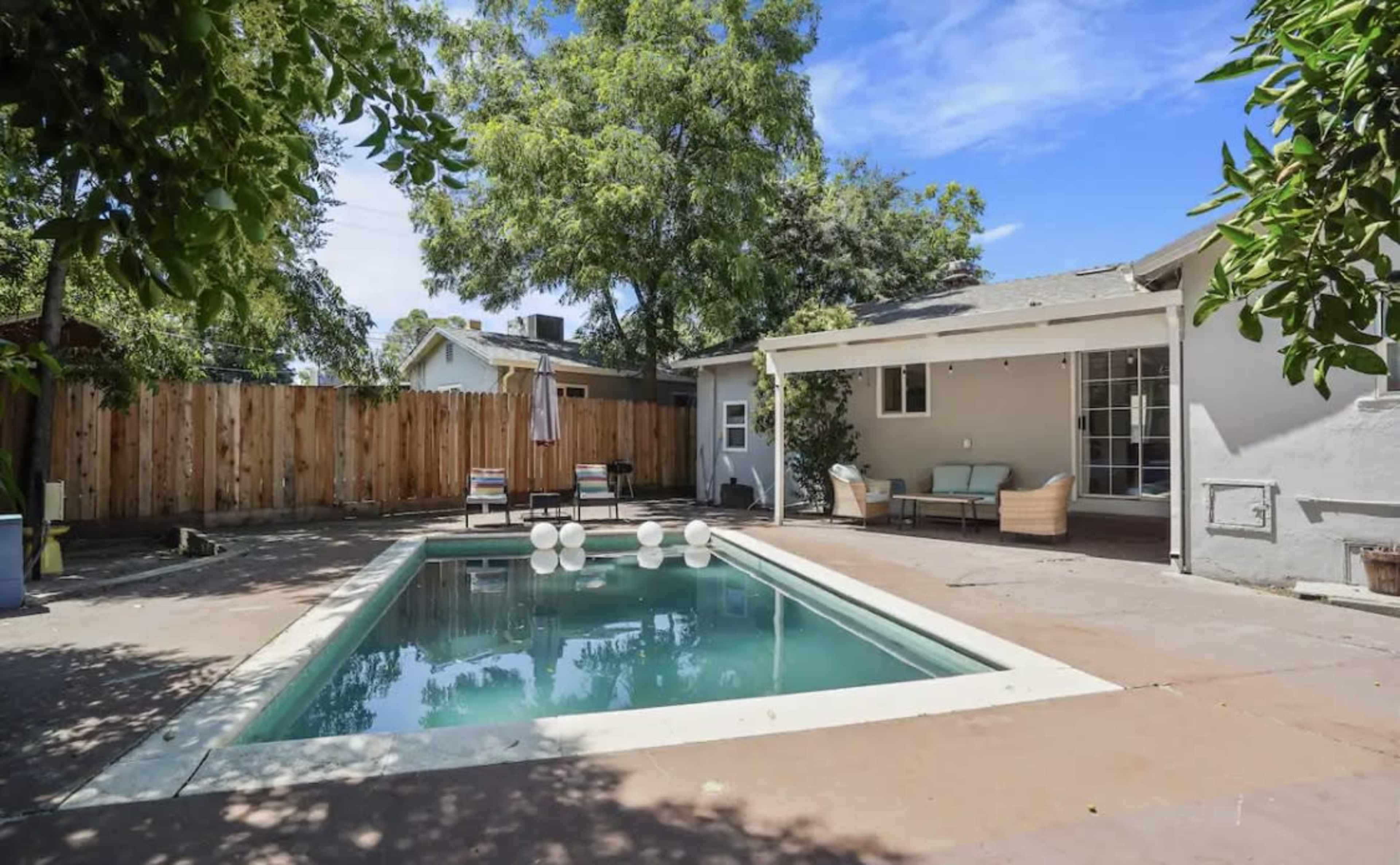 A backyard with a swimming pool surrounded by concrete, patio furniture, and a wooden fence in the background.