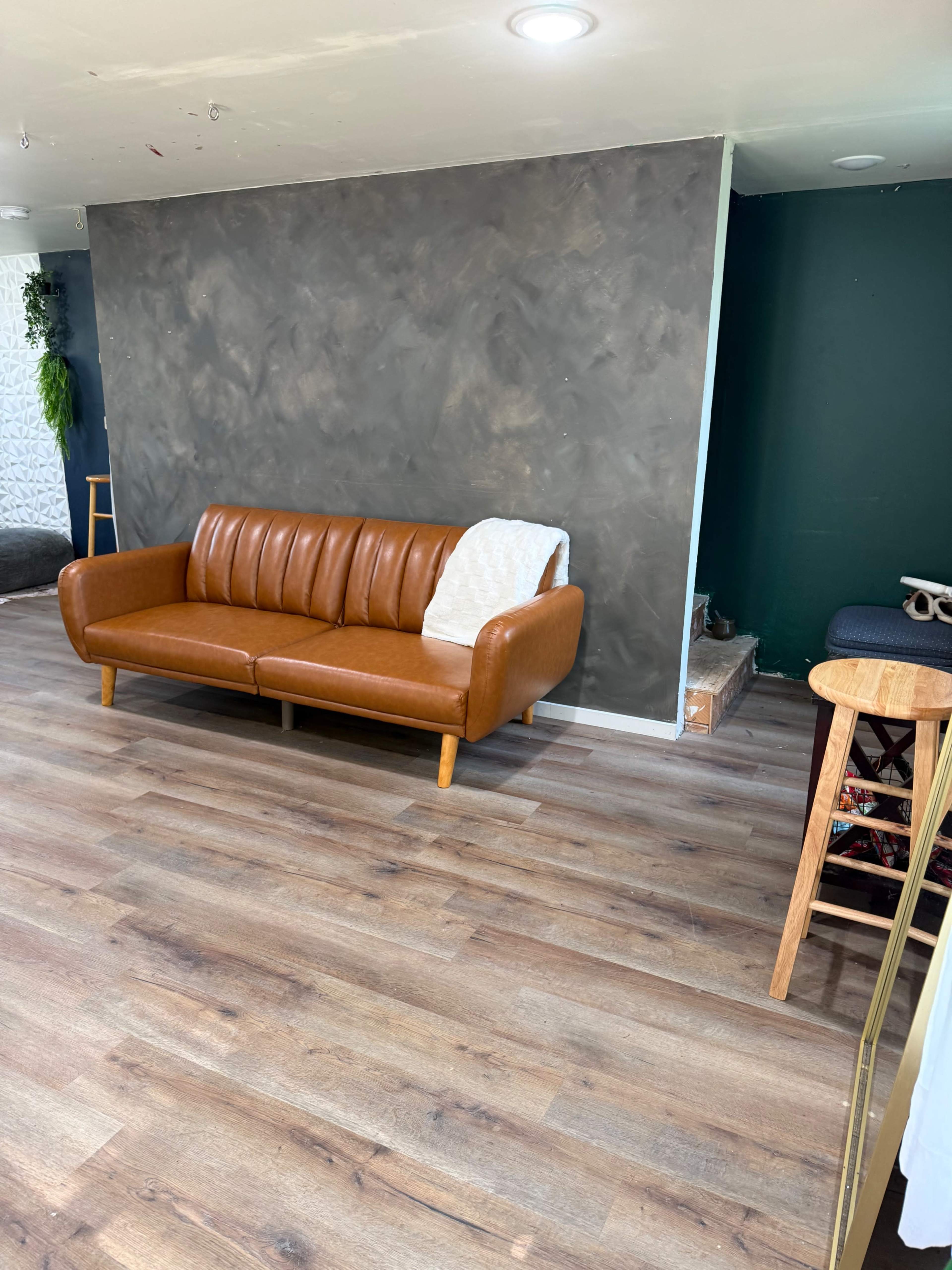 The image shows a living space featuring a brown leather sofa against a textured gray wall, with wooden flooring and some furniture in the background.
