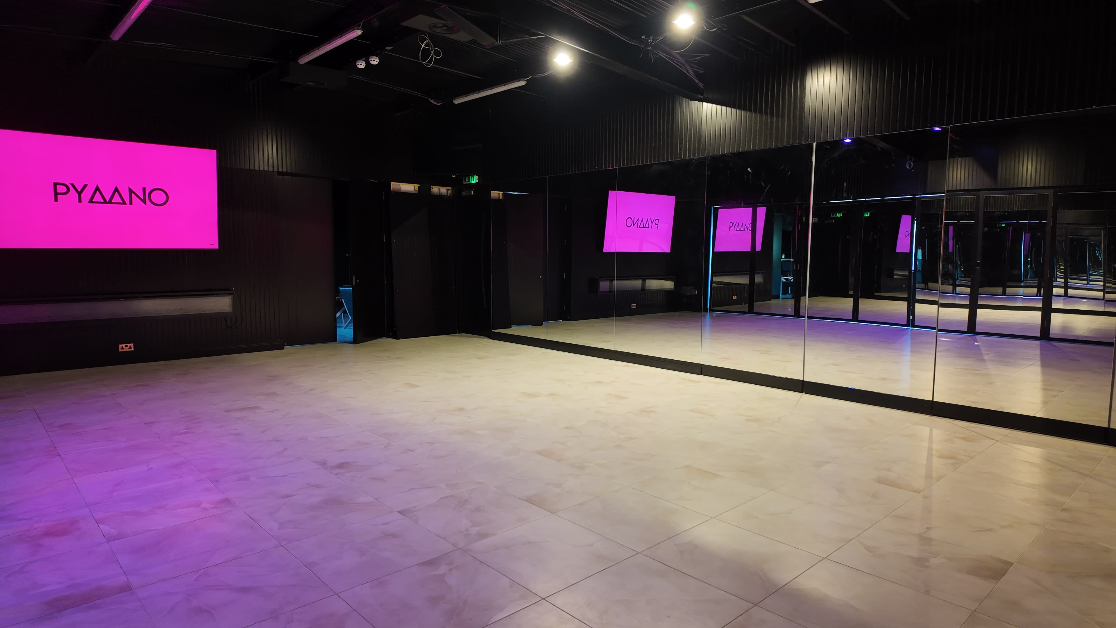 The image shows a spacious, mirrored dance studio with black walls and pink illuminated signage.