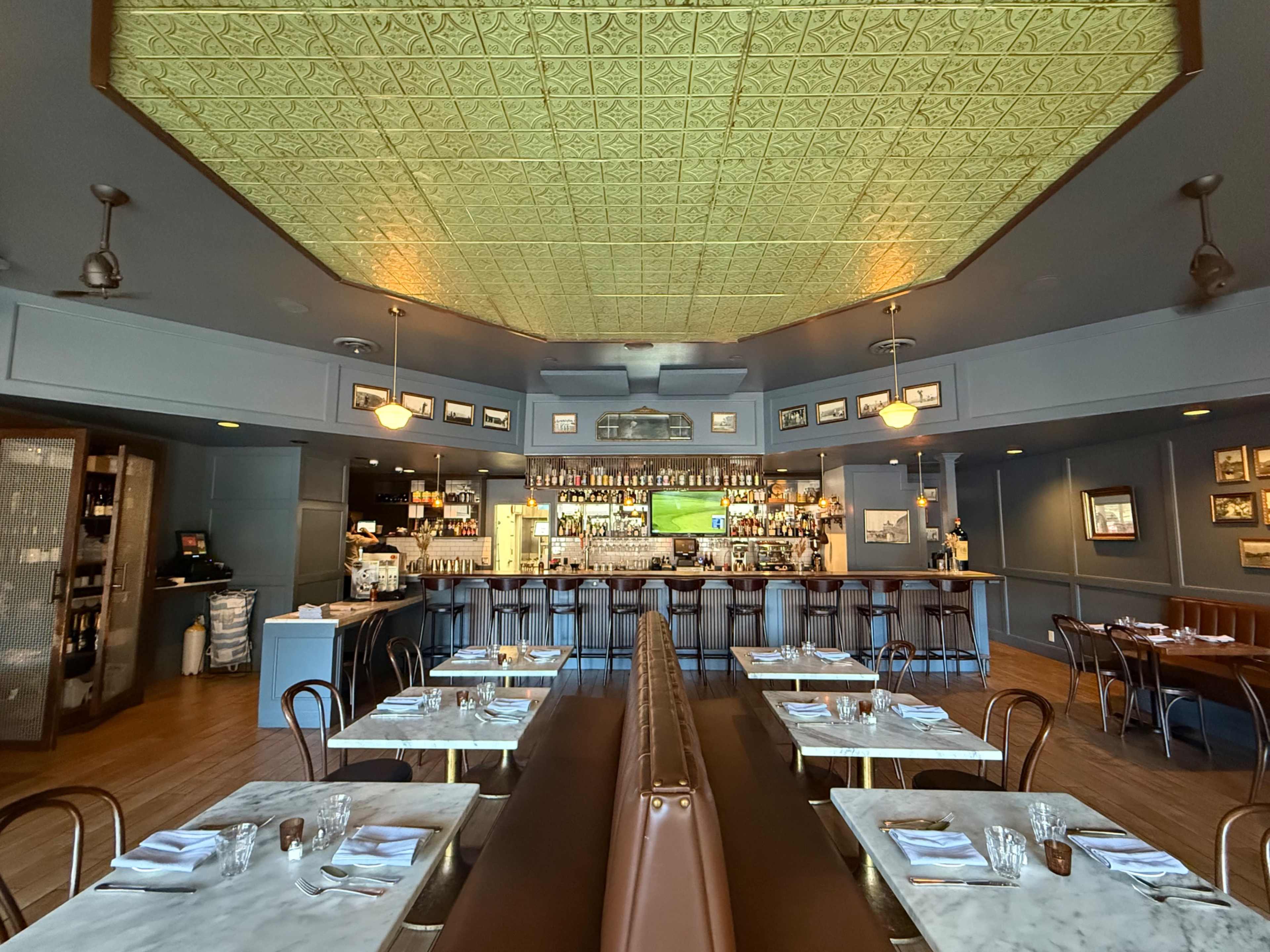 The image shows the interior of a restaurant featuring a bar area, wooden seating, and a decorative ceiling.