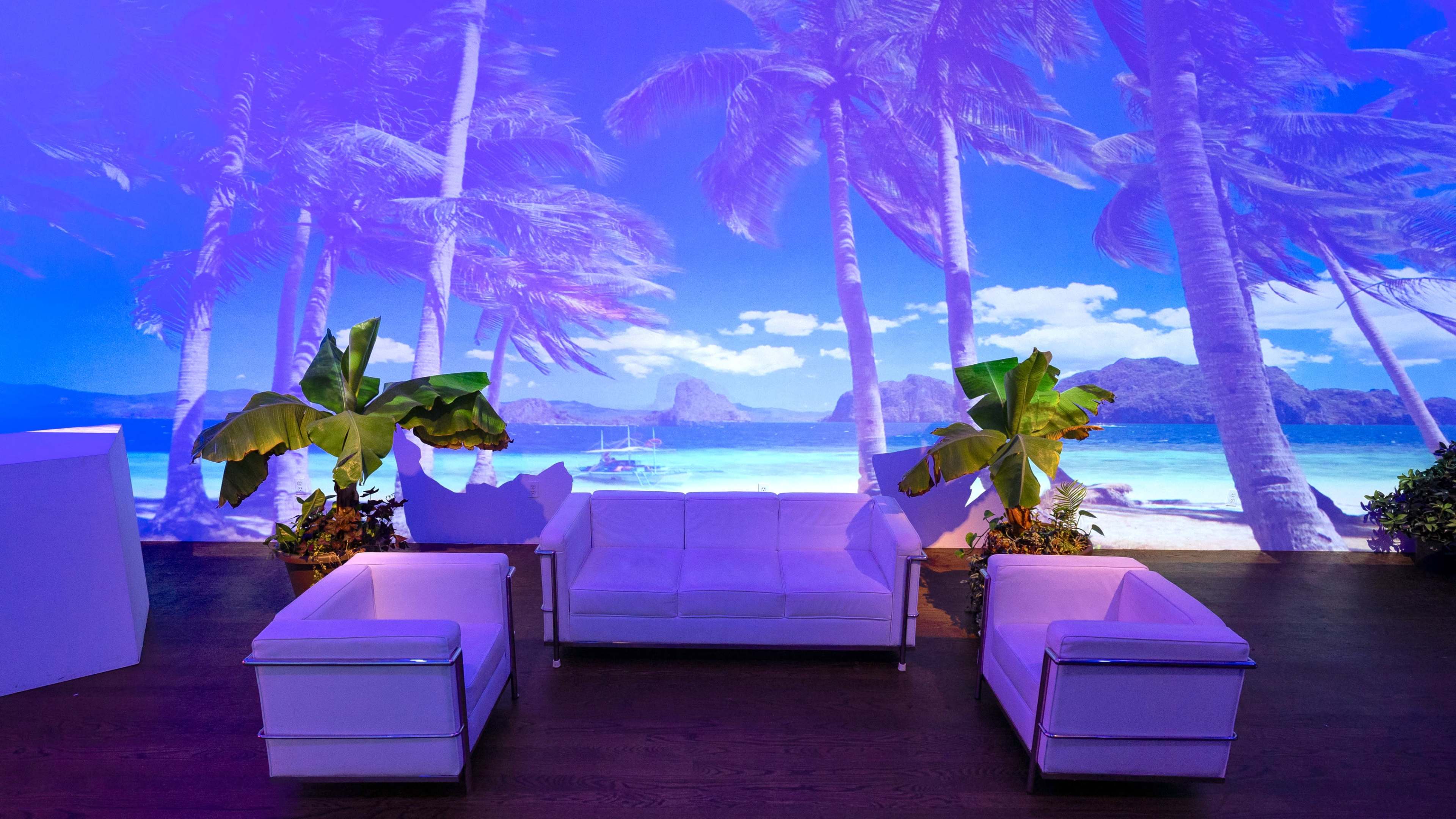 A modern seating area is showcased with a white couch and matching chairs, set against a vibrant beach scene projected on the wall.