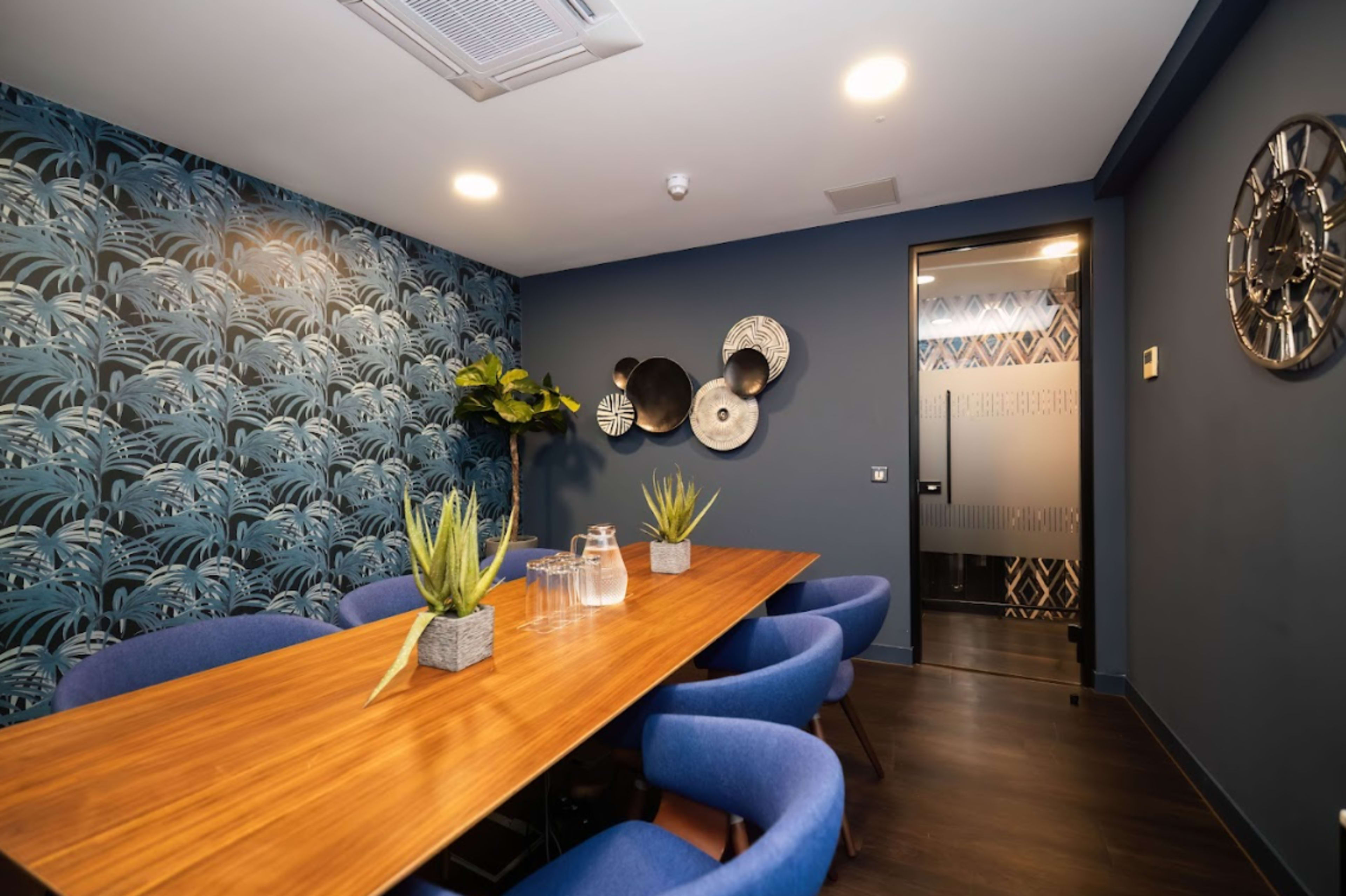 A modern conference room features a long wooden table surrounded by blue chairs, with decorative wall art and plants in a stylish interior.