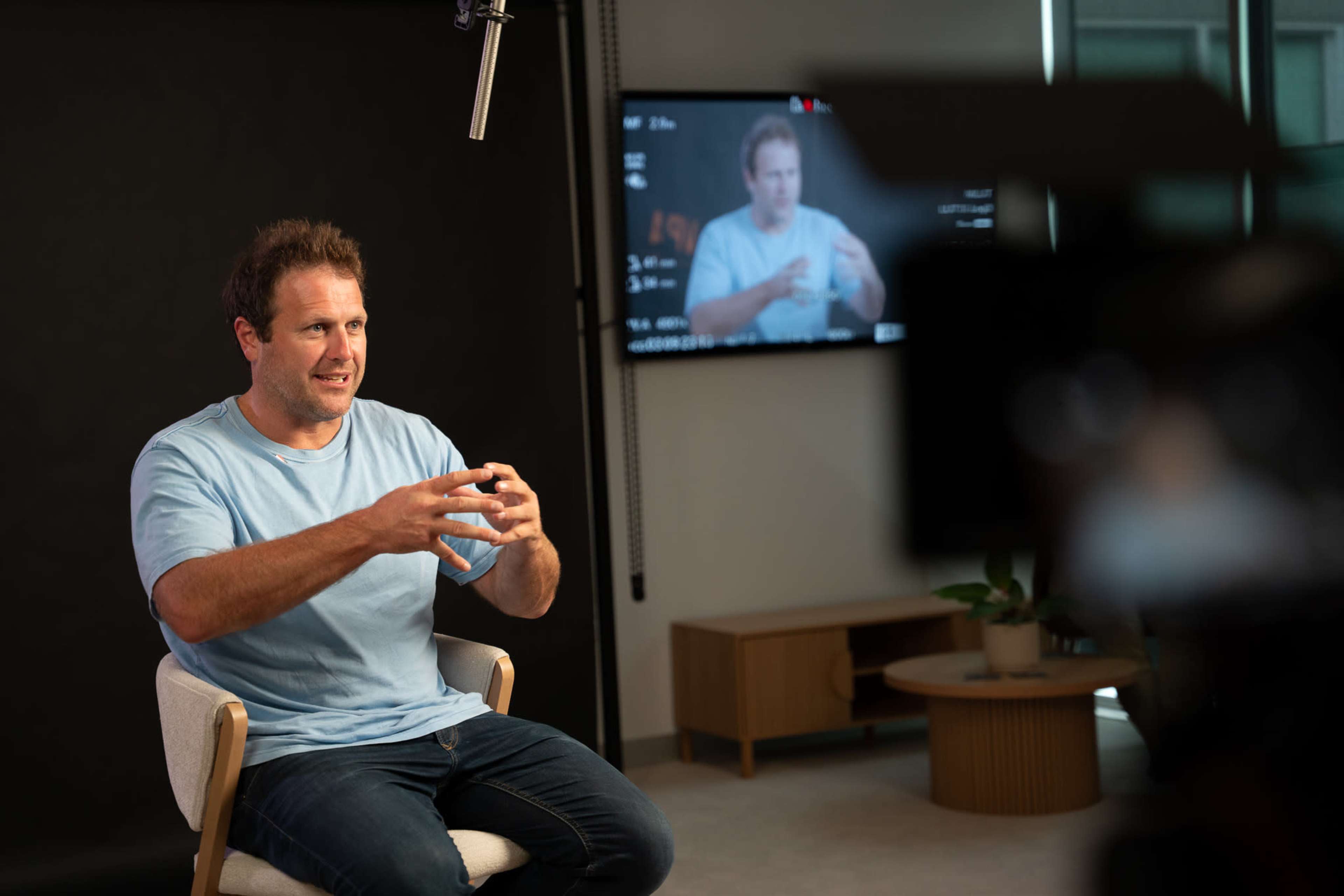 A man in a light blue shirt is sitting in a chair and gesturing while speaking, with a camera focused on him and a monitor displaying his image in the background.