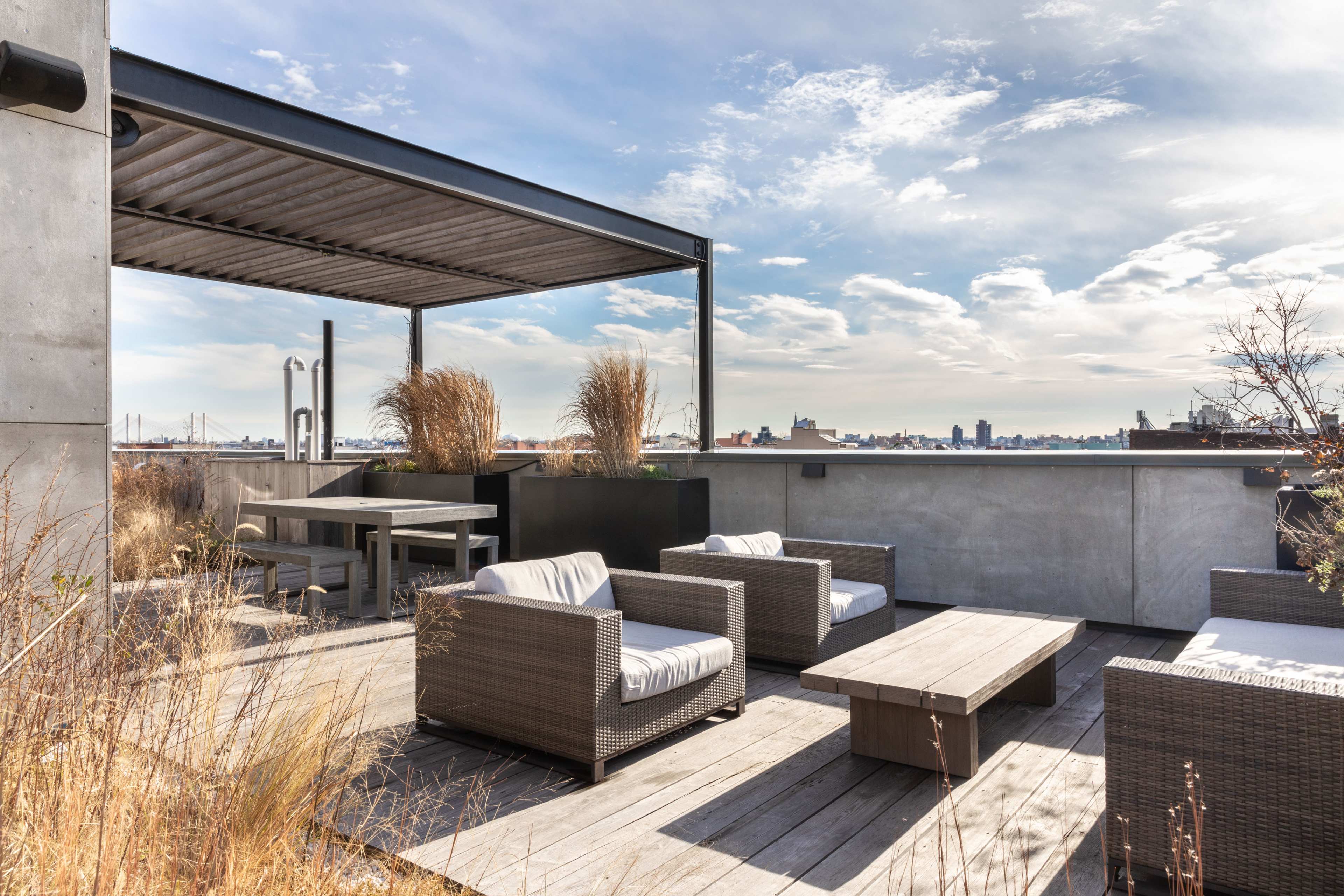 A rooftop terrace featuring outdoor seating and a table under a shaded pergola, with a view of a city skyline in the background.