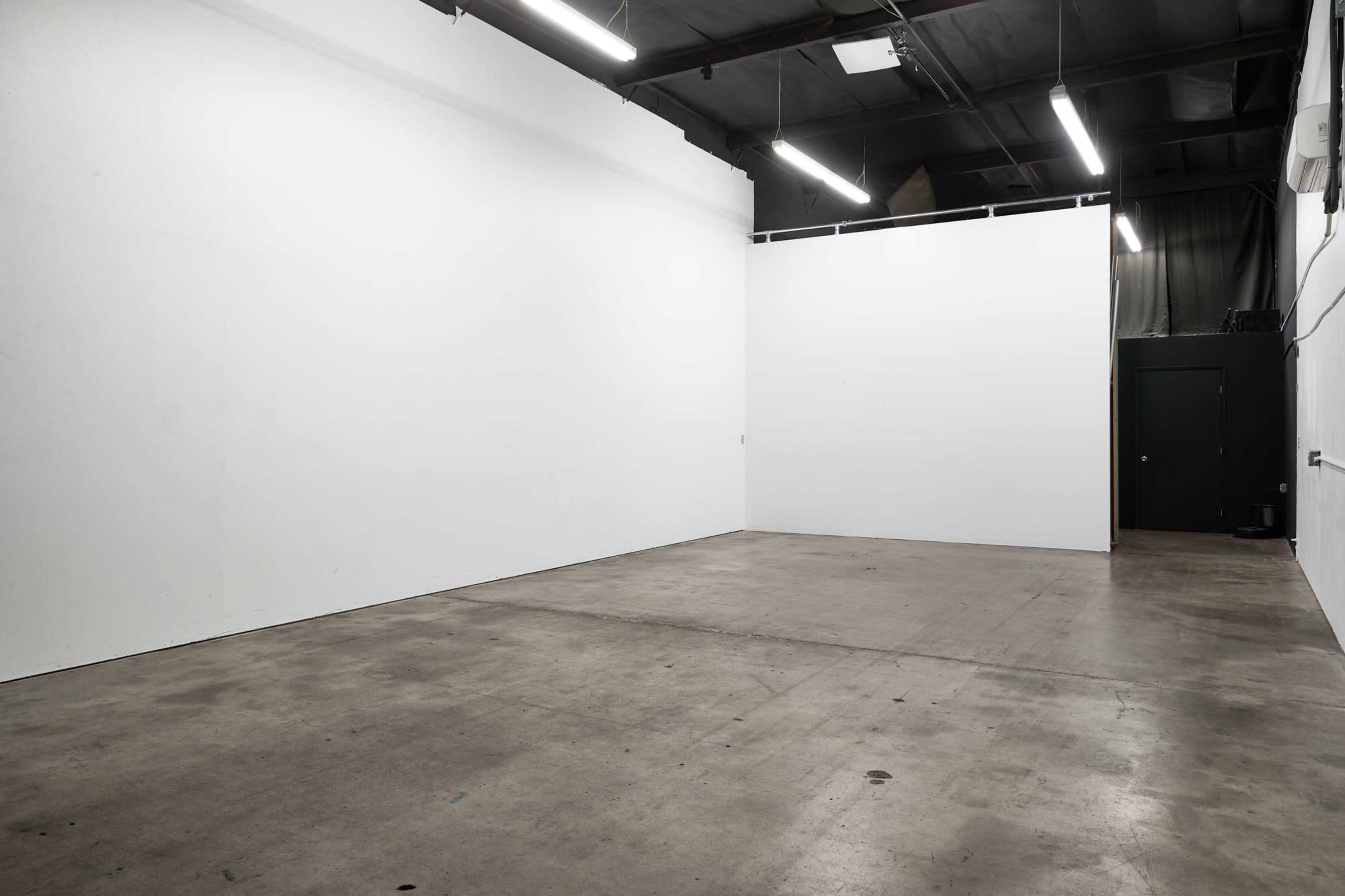 A spacious, empty room with white walls and a concrete floor, illuminated by overhead lights.