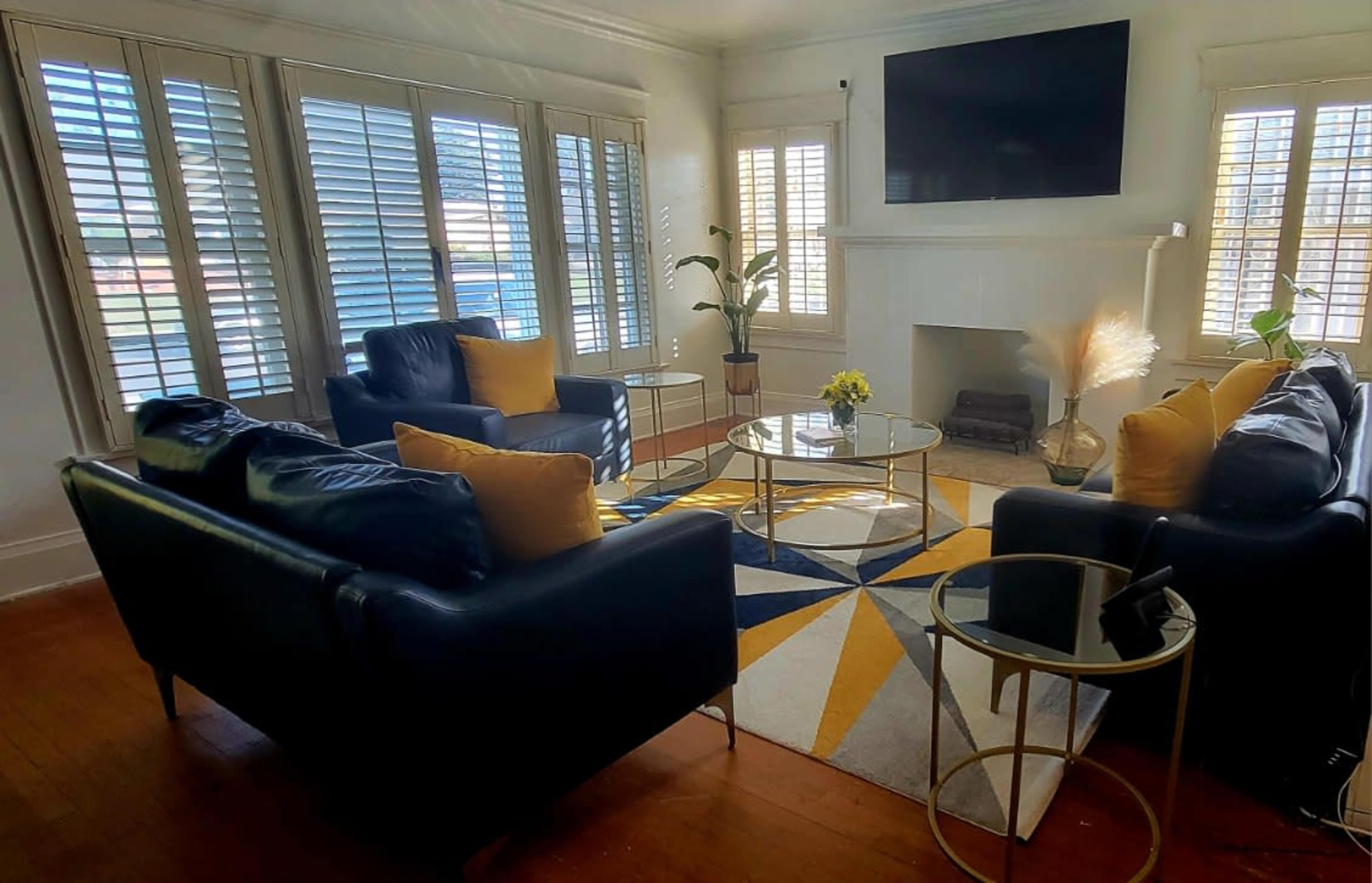 A modern living room features two black sofas with yellow cushions, a glass coffee table, and large windows with wooden shutters.
