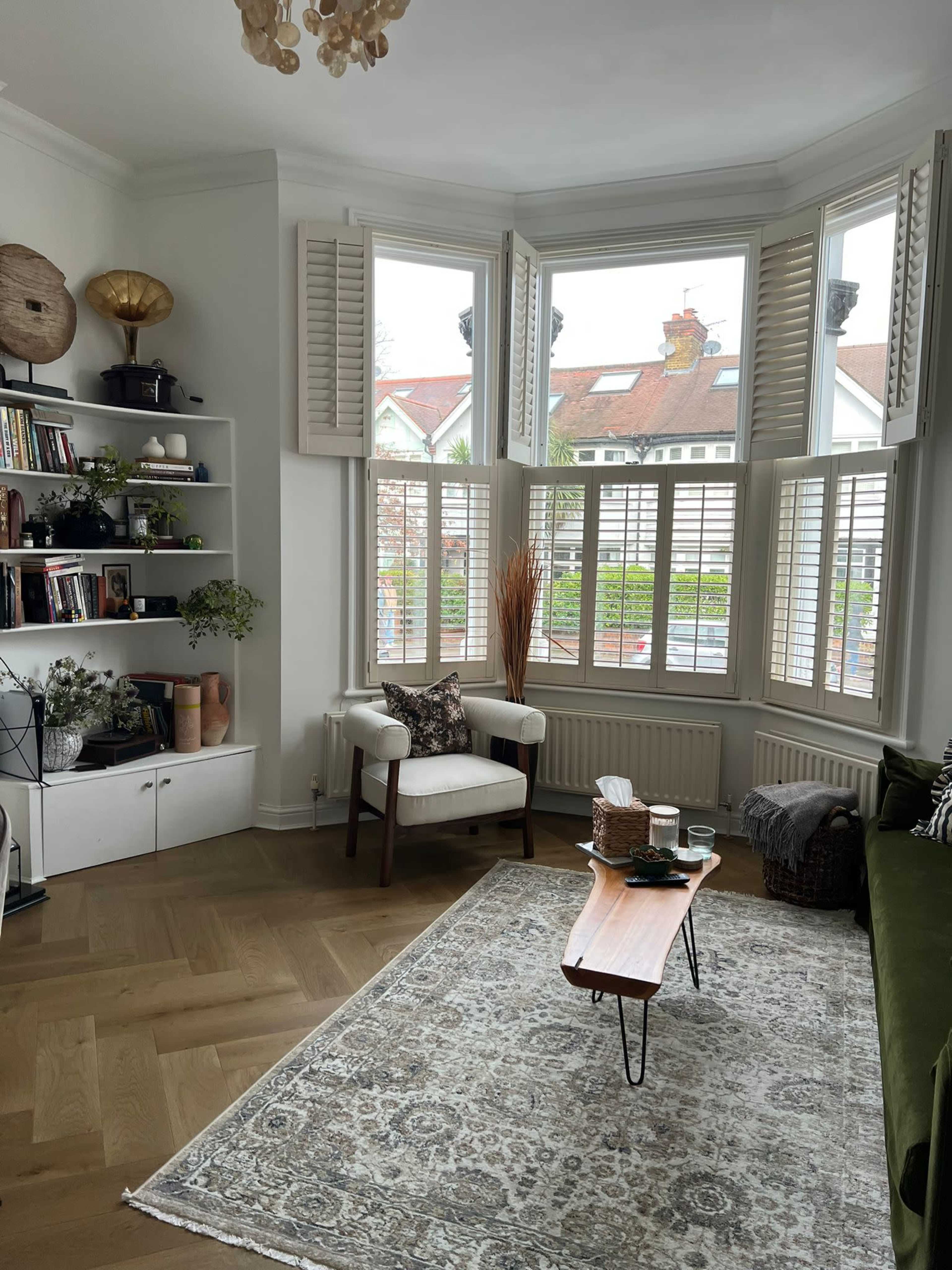 The image shows a cozy living room featuring a green sofa, a wooden coffee table, a bookshelf, and large windows with white shutters.
