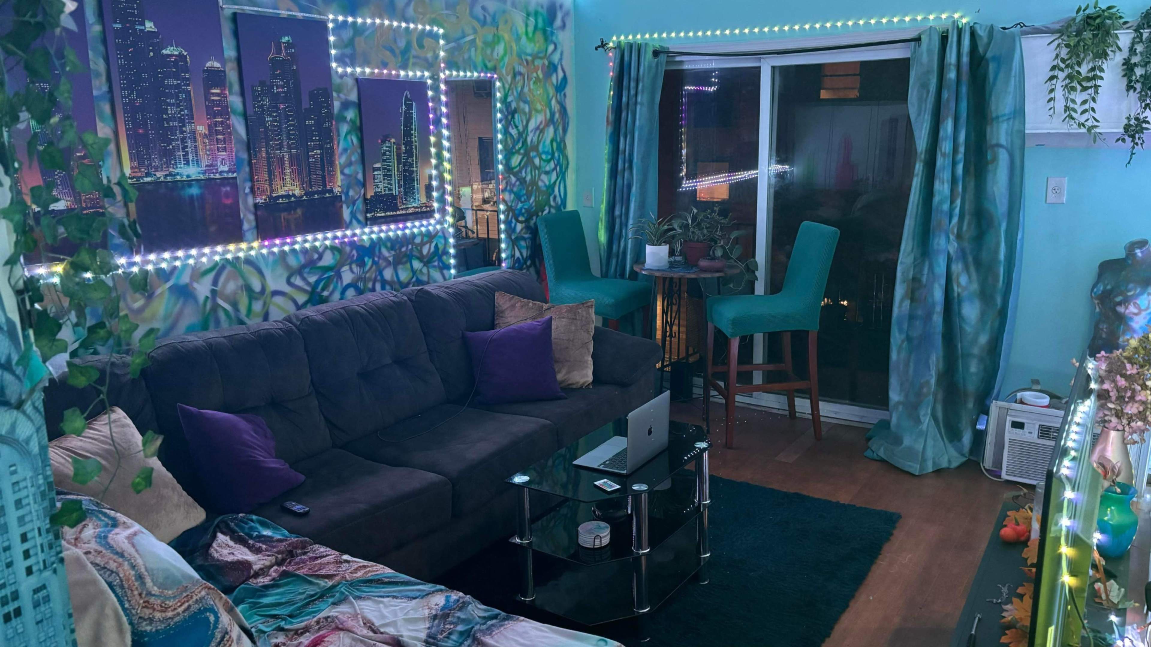The image shows a vibrant living room with a gray sectional sofa, decorative wall art, and a view of a city skyline through a large window.