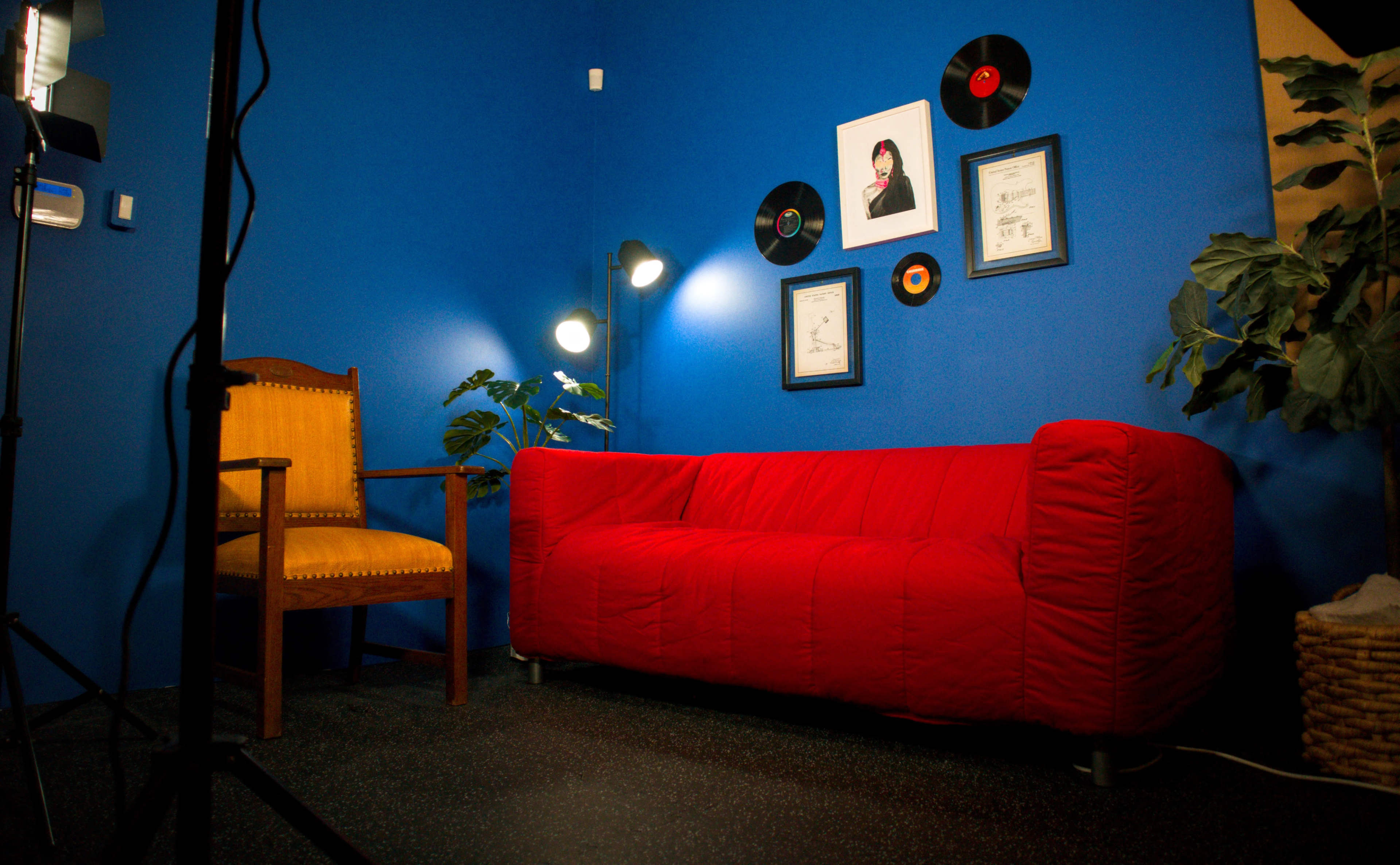 The image shows a bright blue room featuring a red sofa, a wooden chair, and framed records and certificates on the wall, along with a floor lamp illuminating the space.