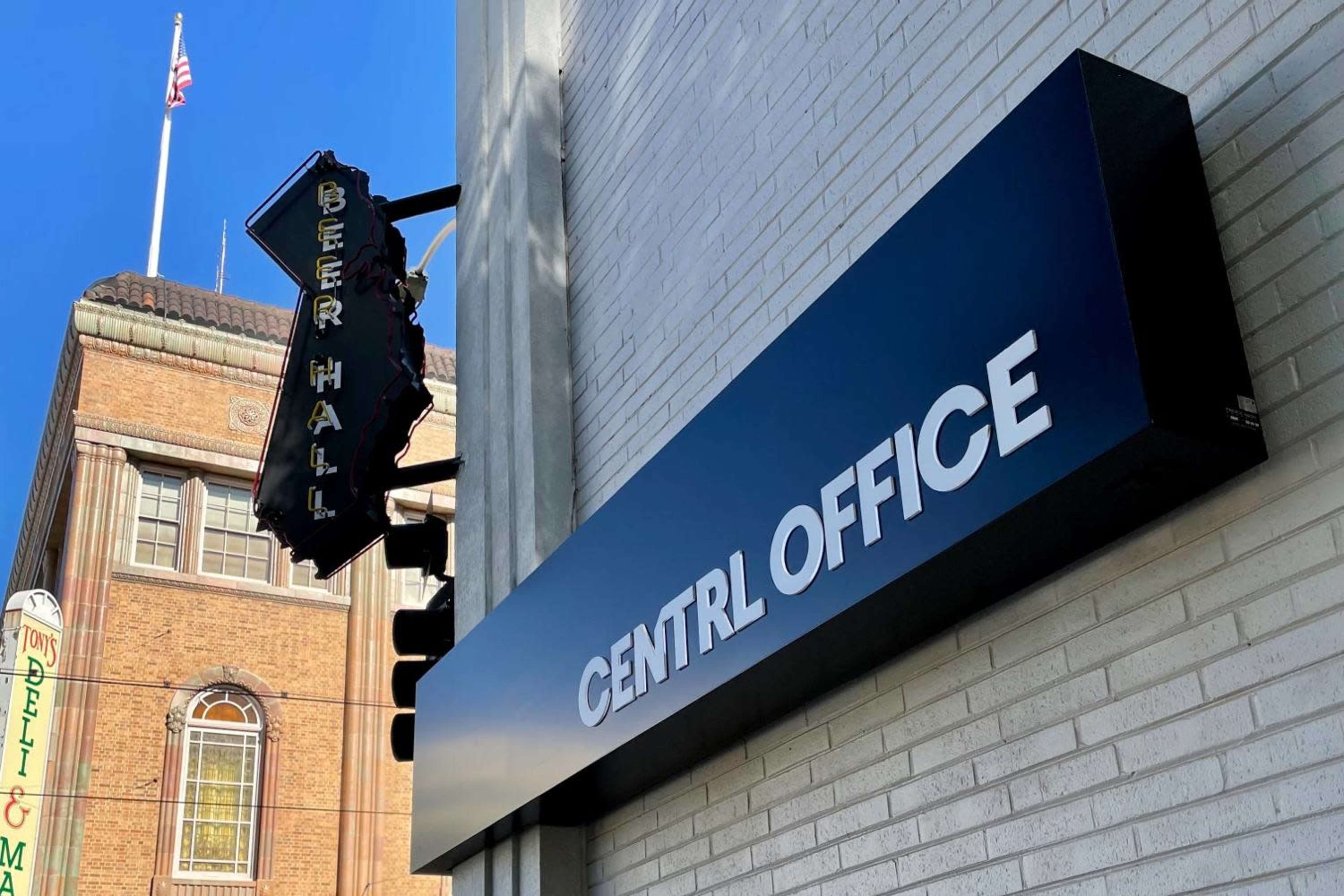 A building facade featuring a sign for a central office, with a beer hall sign partially visible in the background and an American flag atop the nearby structure.