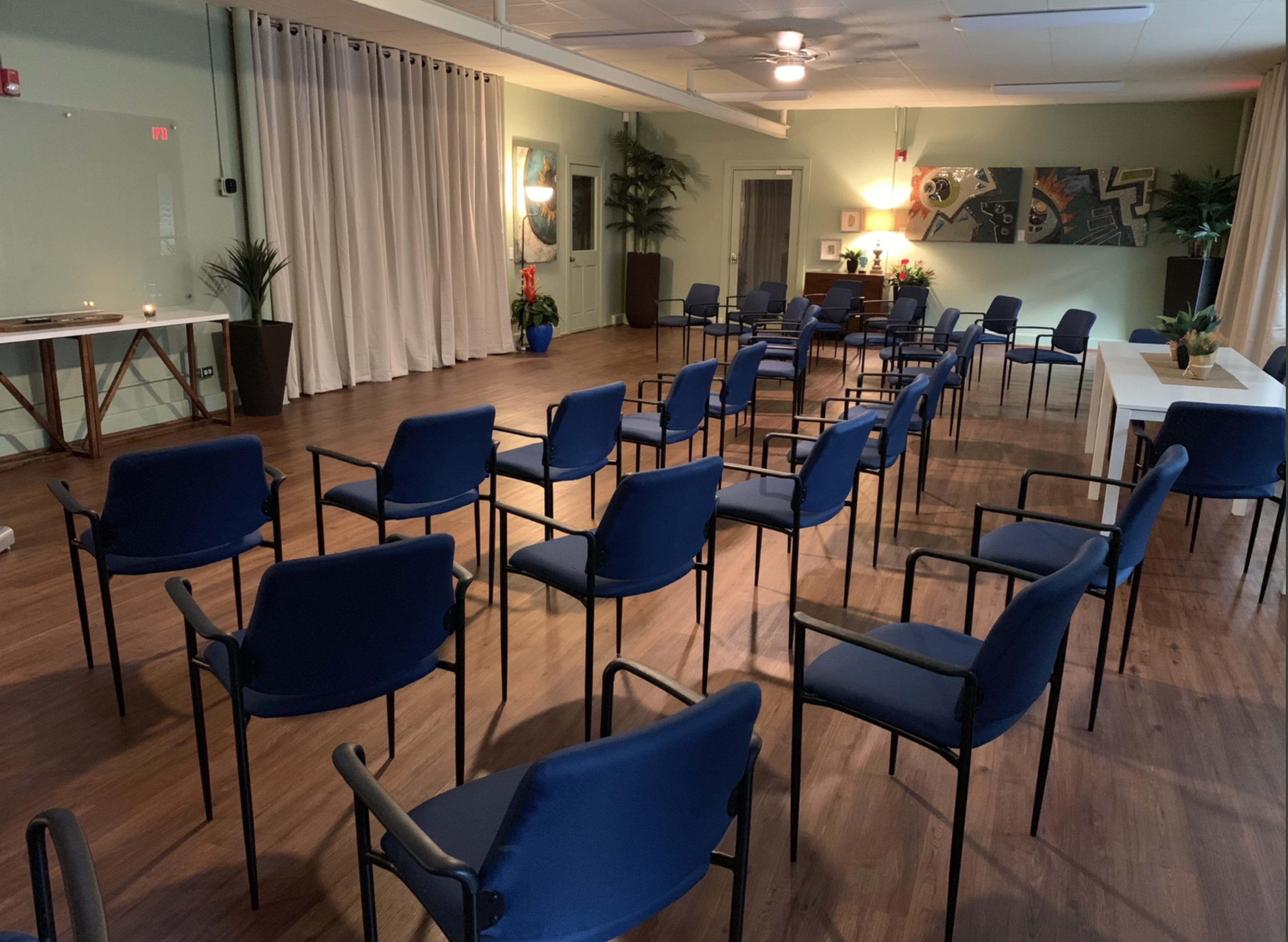 A spacious room with several blue chairs arranged in a circular pattern, surrounded by plants and artwork on the walls.