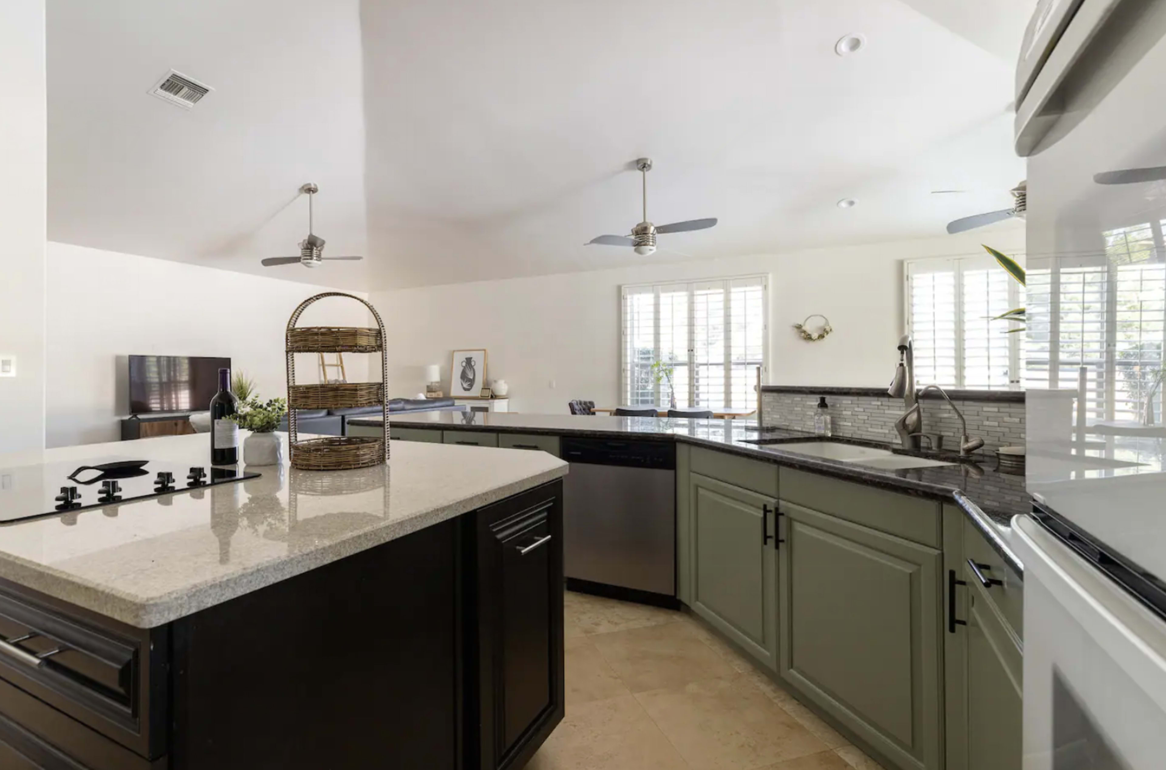 A modern kitchen featuring an island with granite countertops, stainless steel appliances, and large windows with plantation shutters.