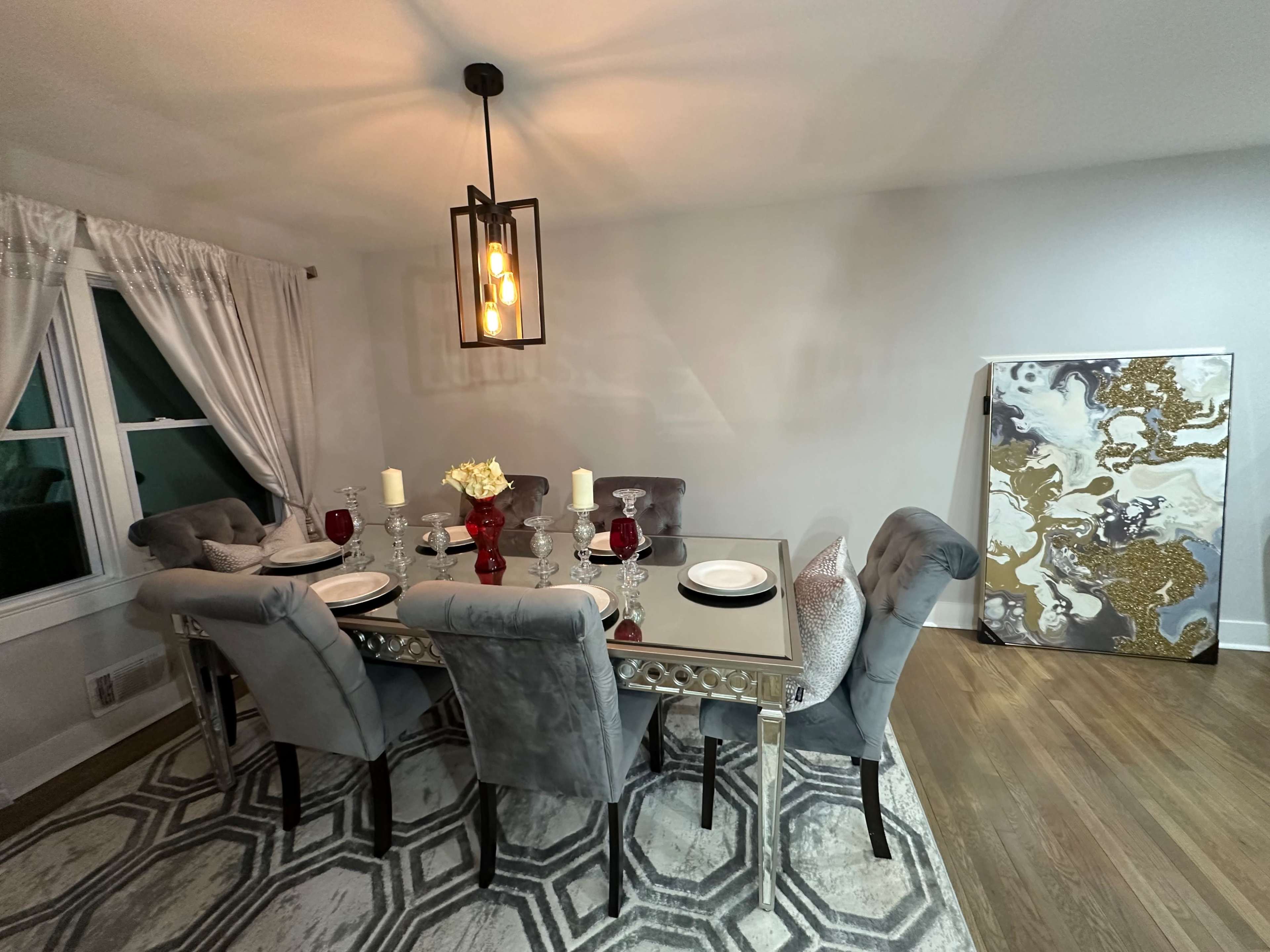 A modern dining area features a mirrored table set with dishes and red glasses, surrounded by plush gray chairs, with abstract art displayed on the wall.