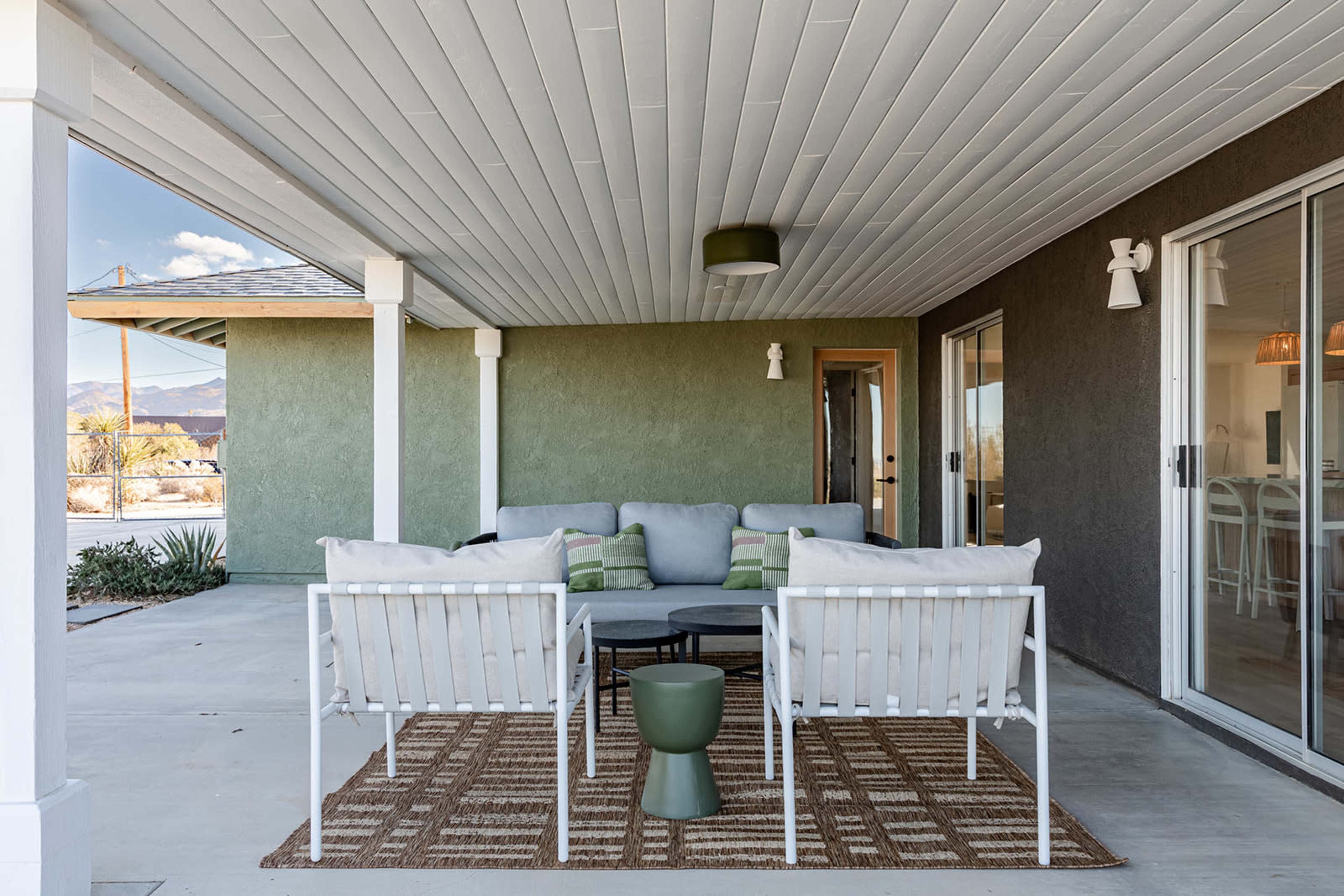 The image shows a covered outdoor seating area featuring a gray sectional sofa, two white armchairs, and a round black table on a patterned rug.