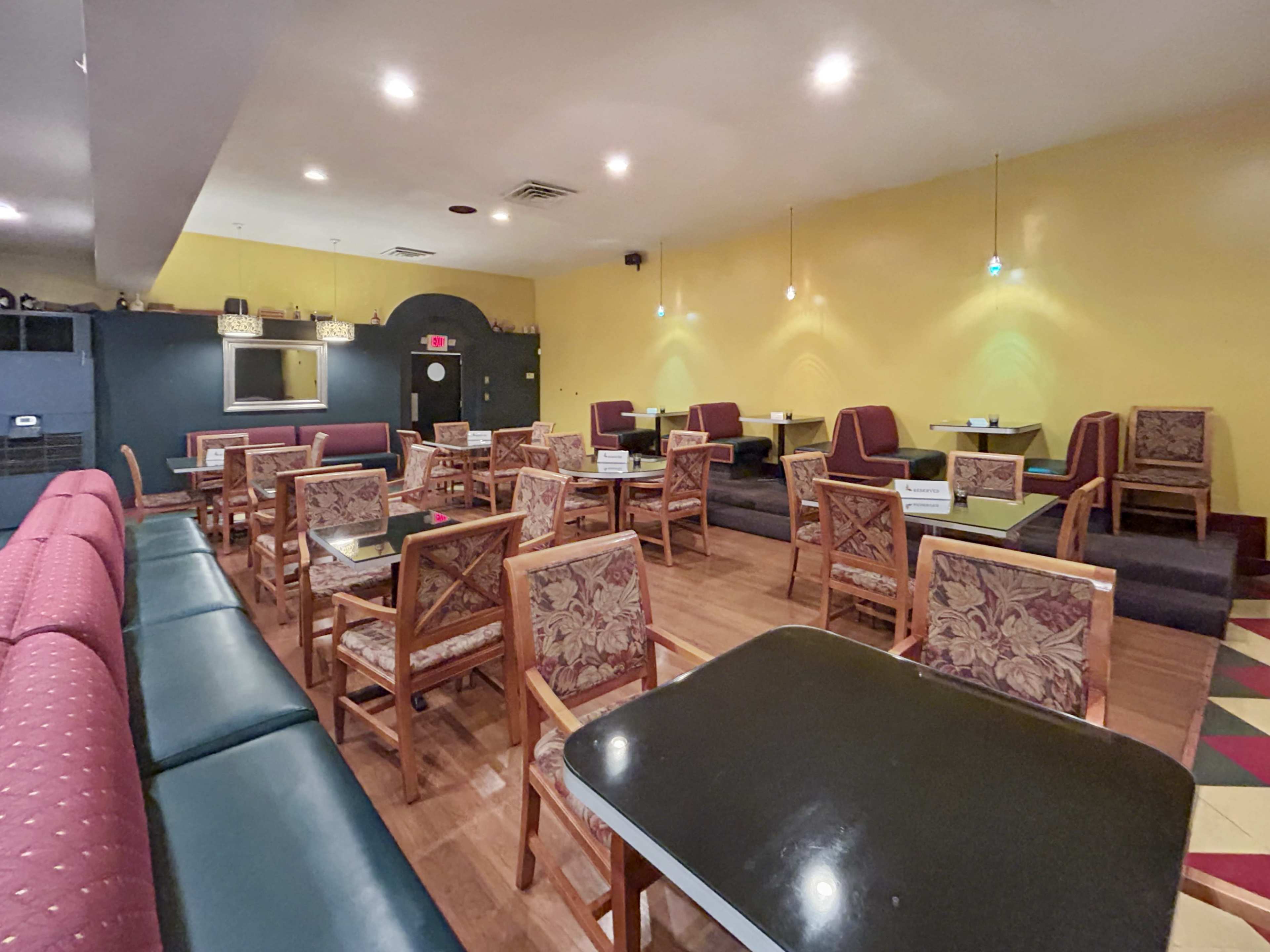 An empty cafe with wooden tables and chairs arranged on a patterned floor, and yellow walls adorned with wall lamps.