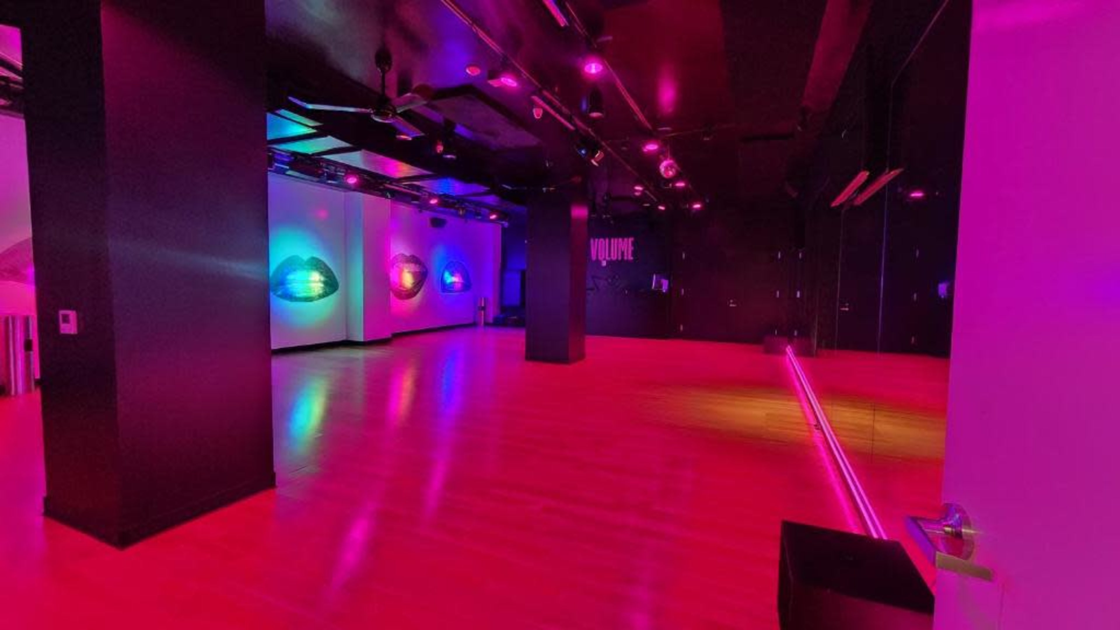 The image shows a spacious empty room with glossy wooden floors, illuminated by colorful pink and blue lights, and featuring abstract wall art.