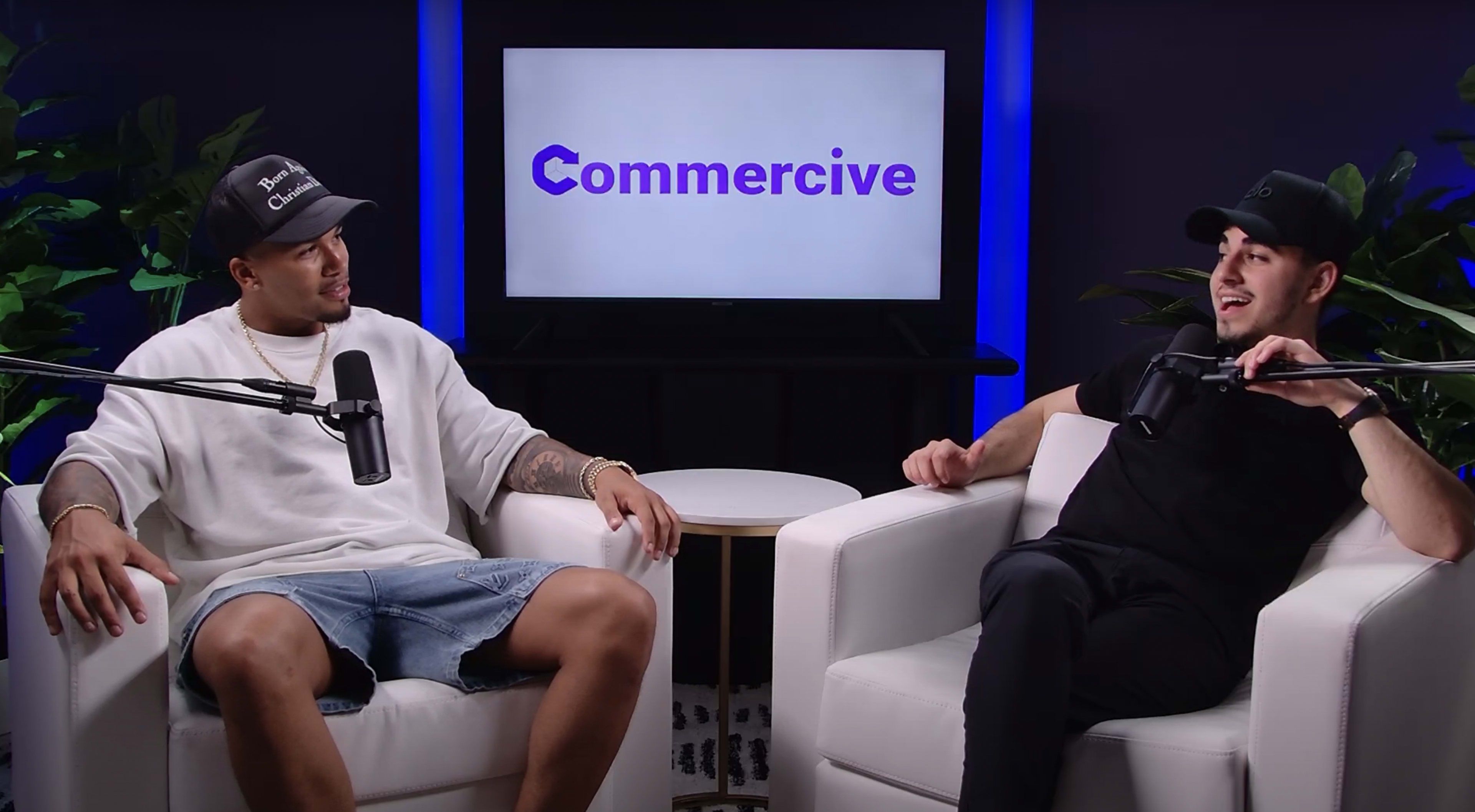 Two individuals are seated in white armchairs in front of a screen displaying the word "Commercial."