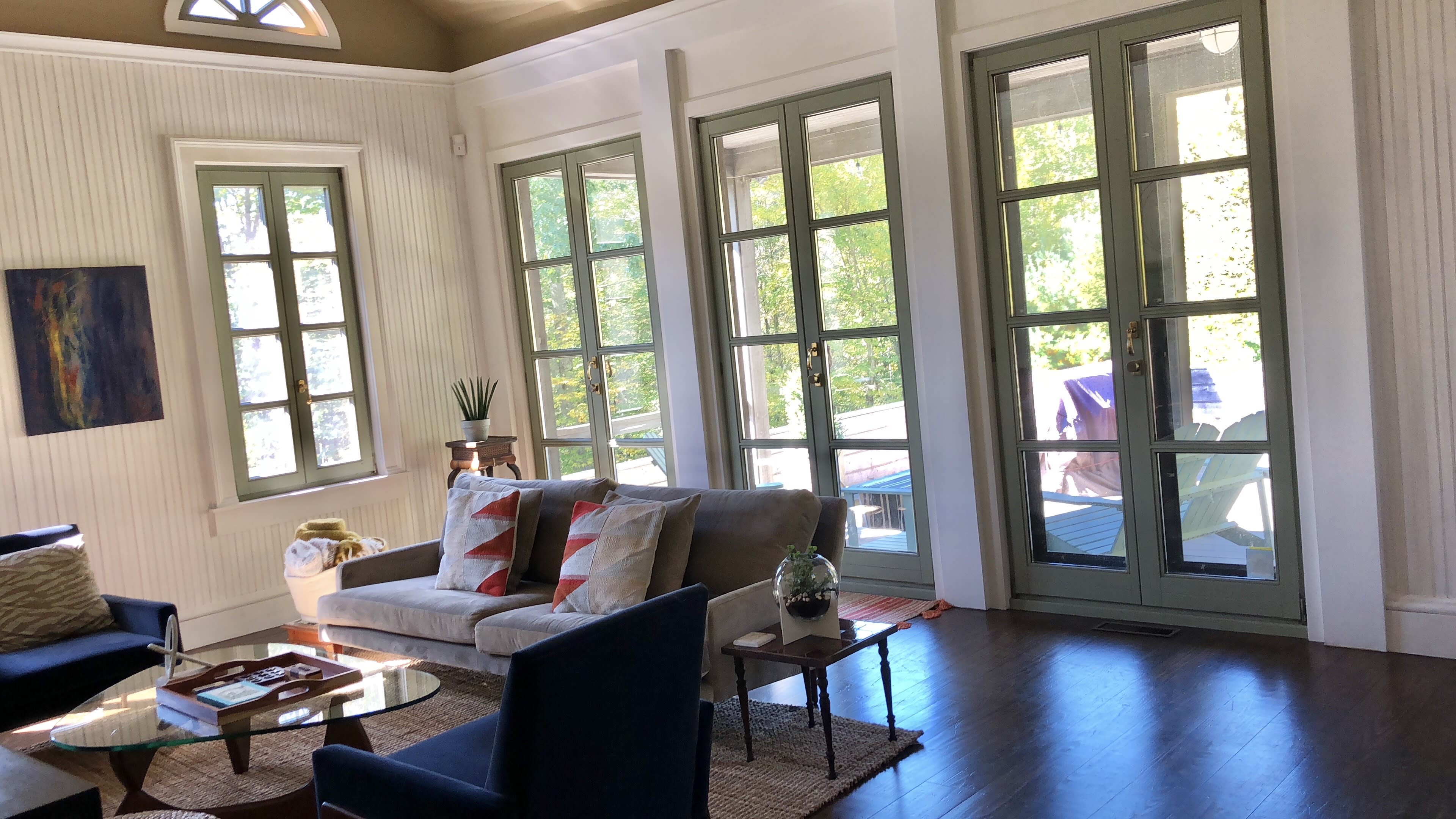 The room features a light-colored sofa adorned with cushions, several large windows, and a coffee table on a wooden floor.
