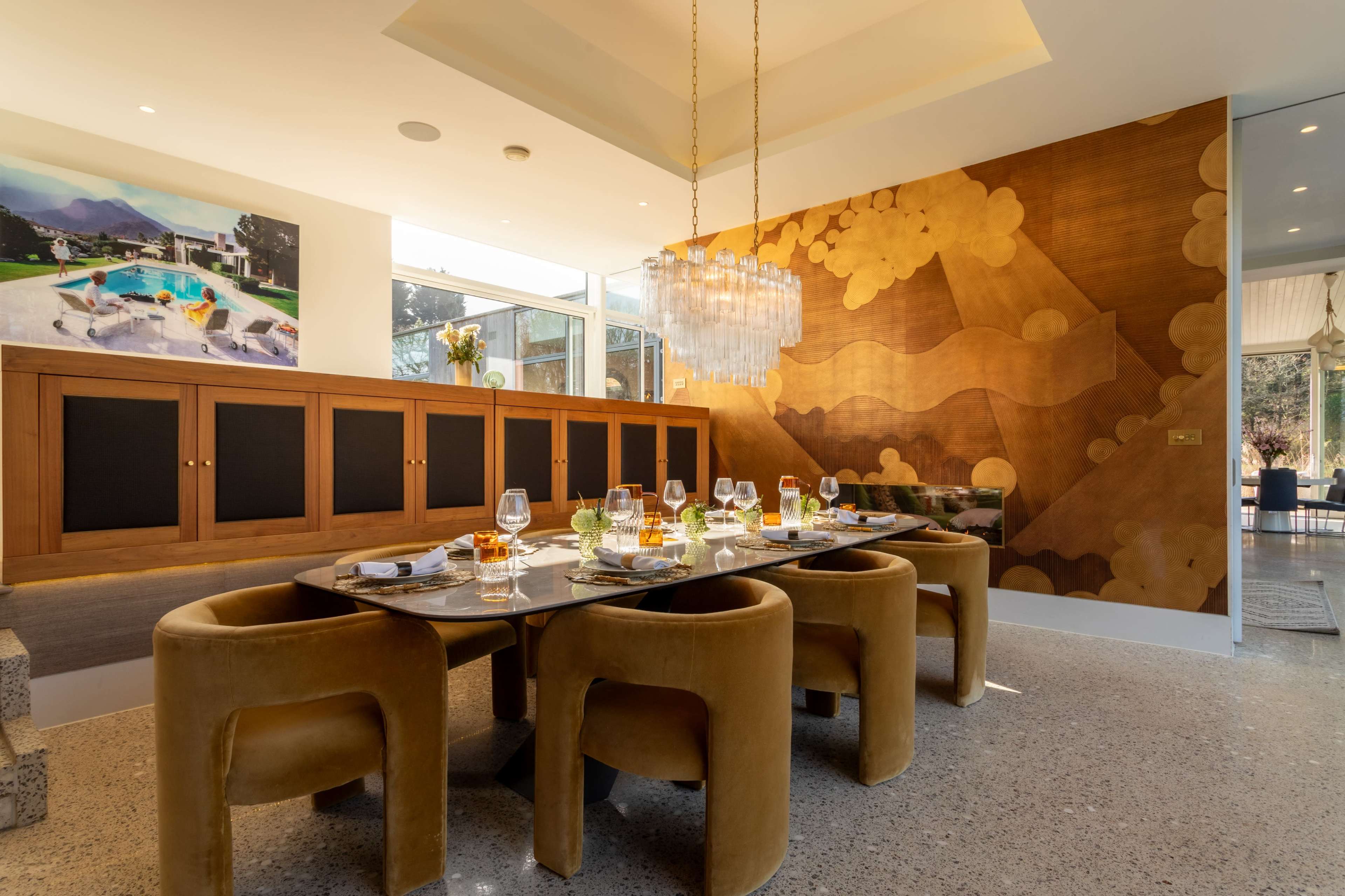 The image shows a modern dining room featuring a large table set for a meal, surrounded by rounded chairs, with an artistic wall mural and a large window bringing in natural light.