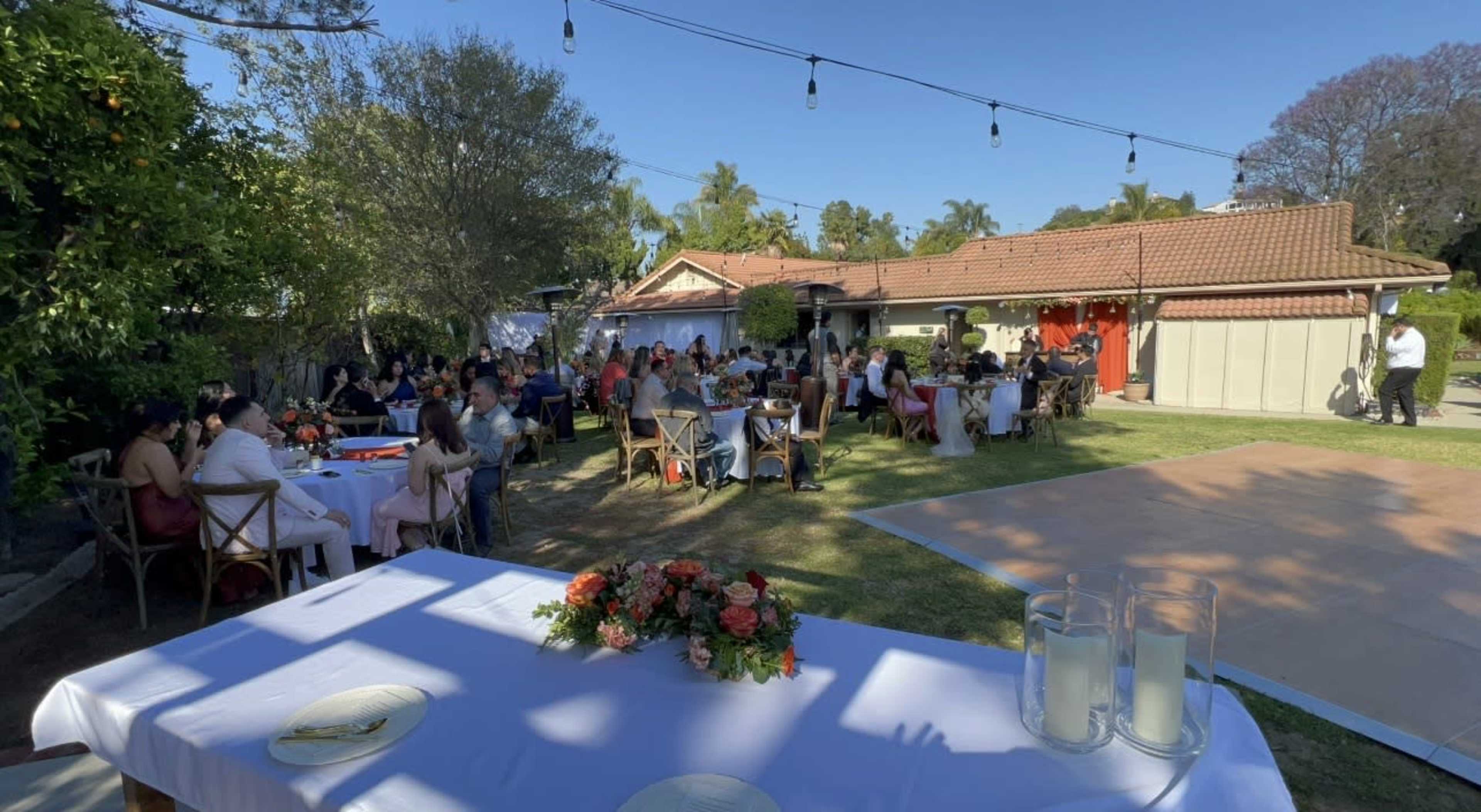 A garden party is taking place, with tables set for dining and a group of guests enjoying the outdoor setting.