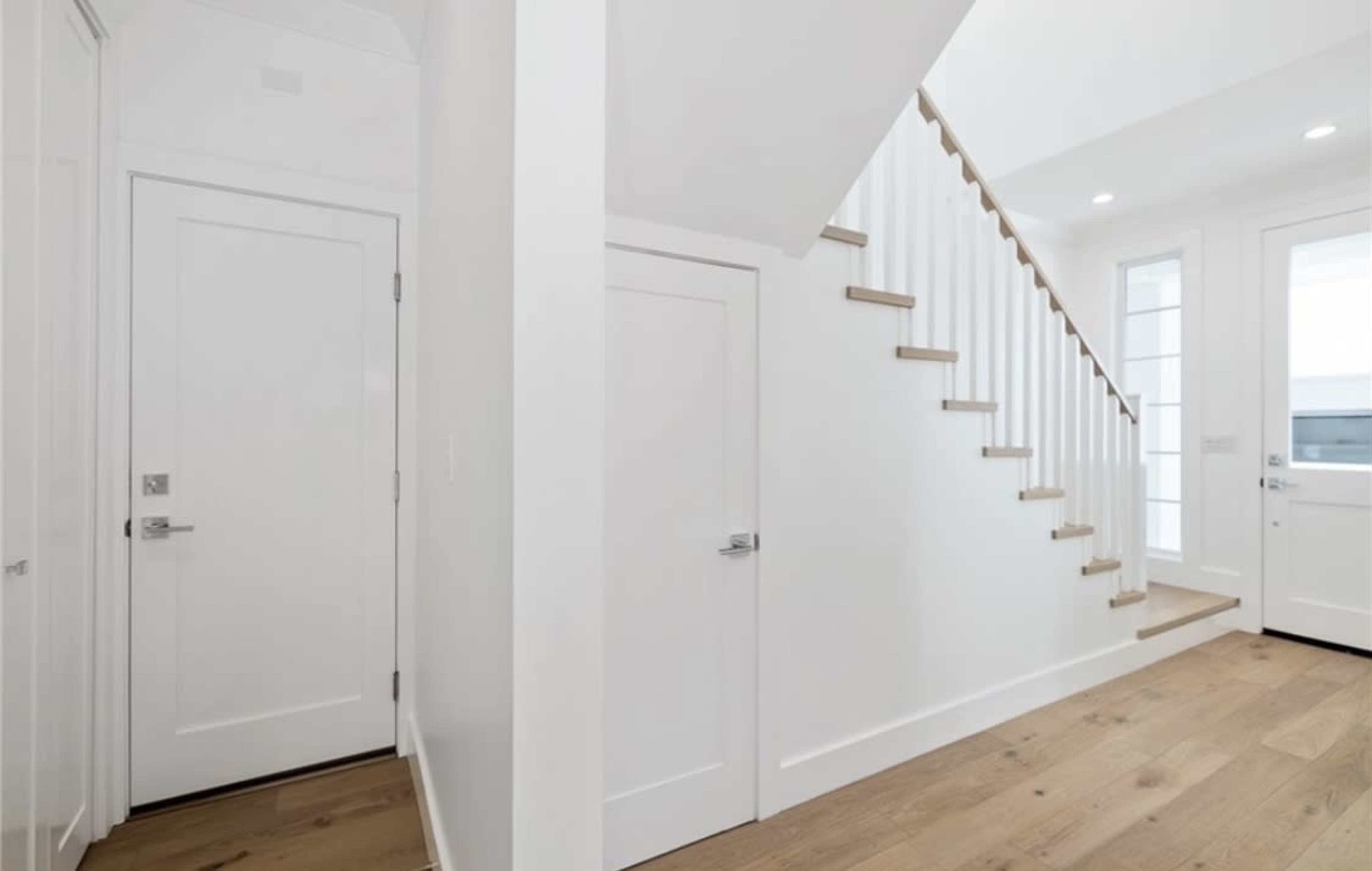 A white hallway features a staircase, two closed doors, and a large window providing natural light.