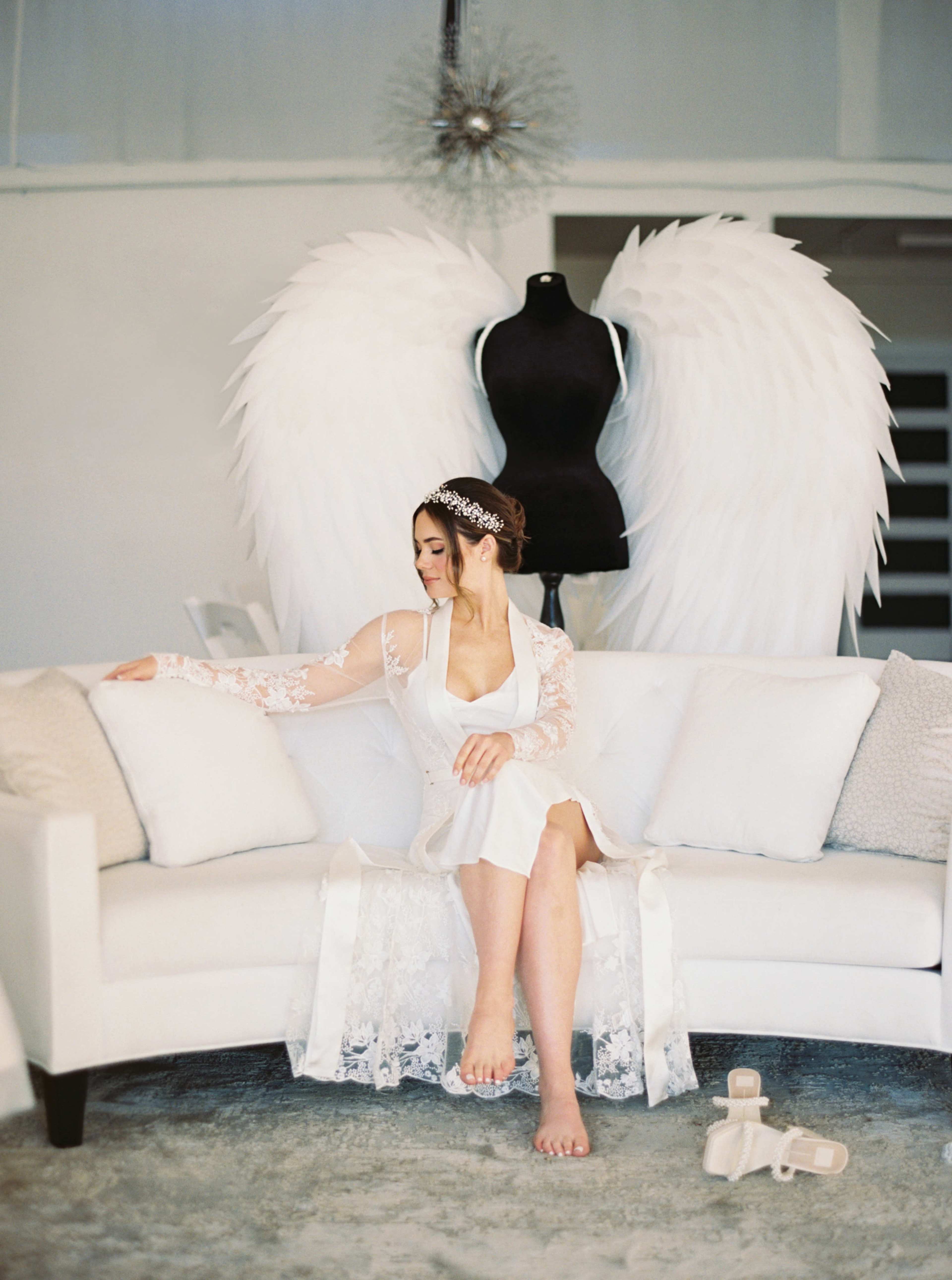 A woman in a white dress sits on a sofa in front of a mannequin with large, white angel wings.