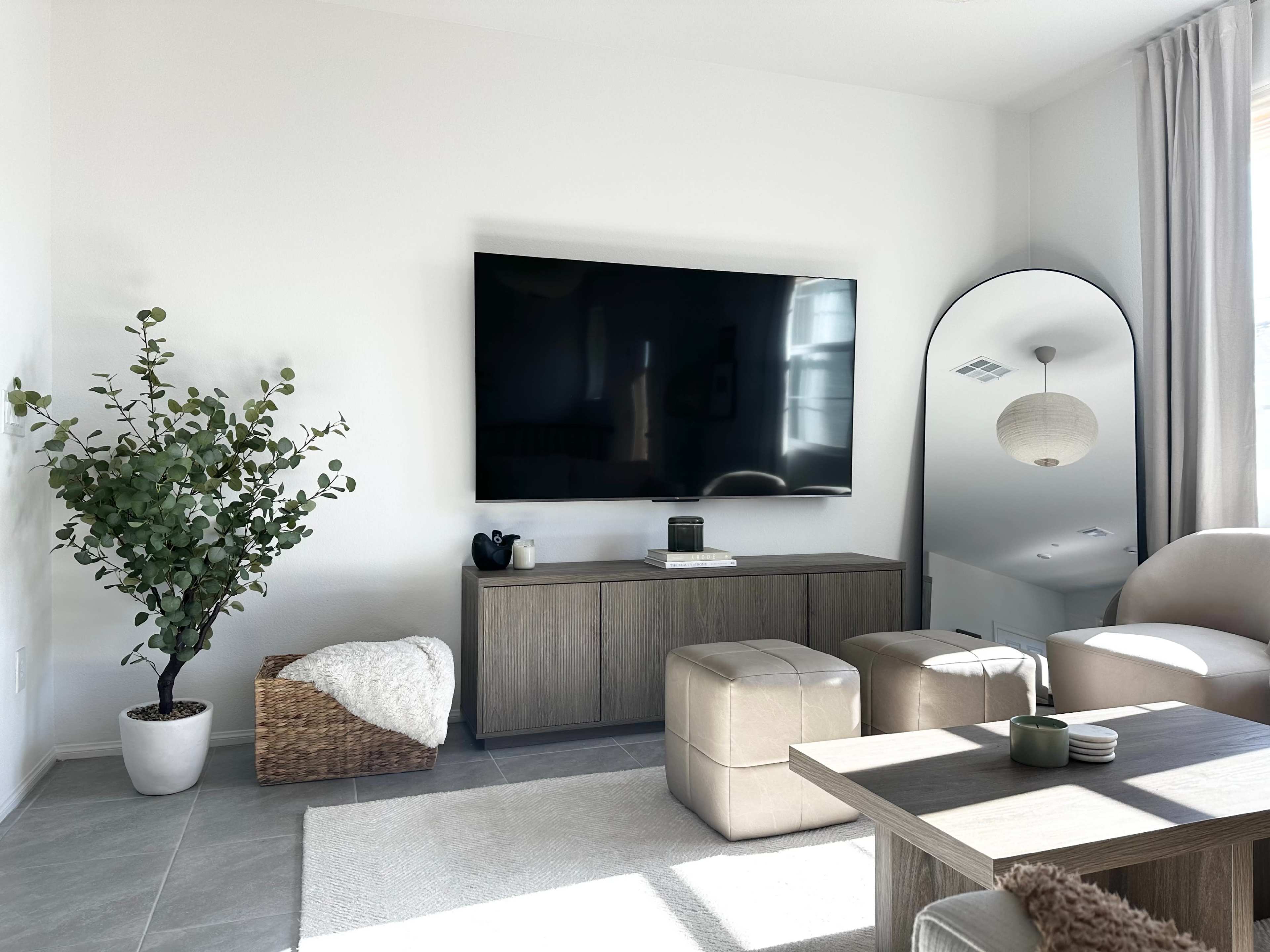 The living room features a large wall-mounted TV above a wooden console, paired with a round mirror, potted plant, and upholstered ottomans arranged around a coffee table.
