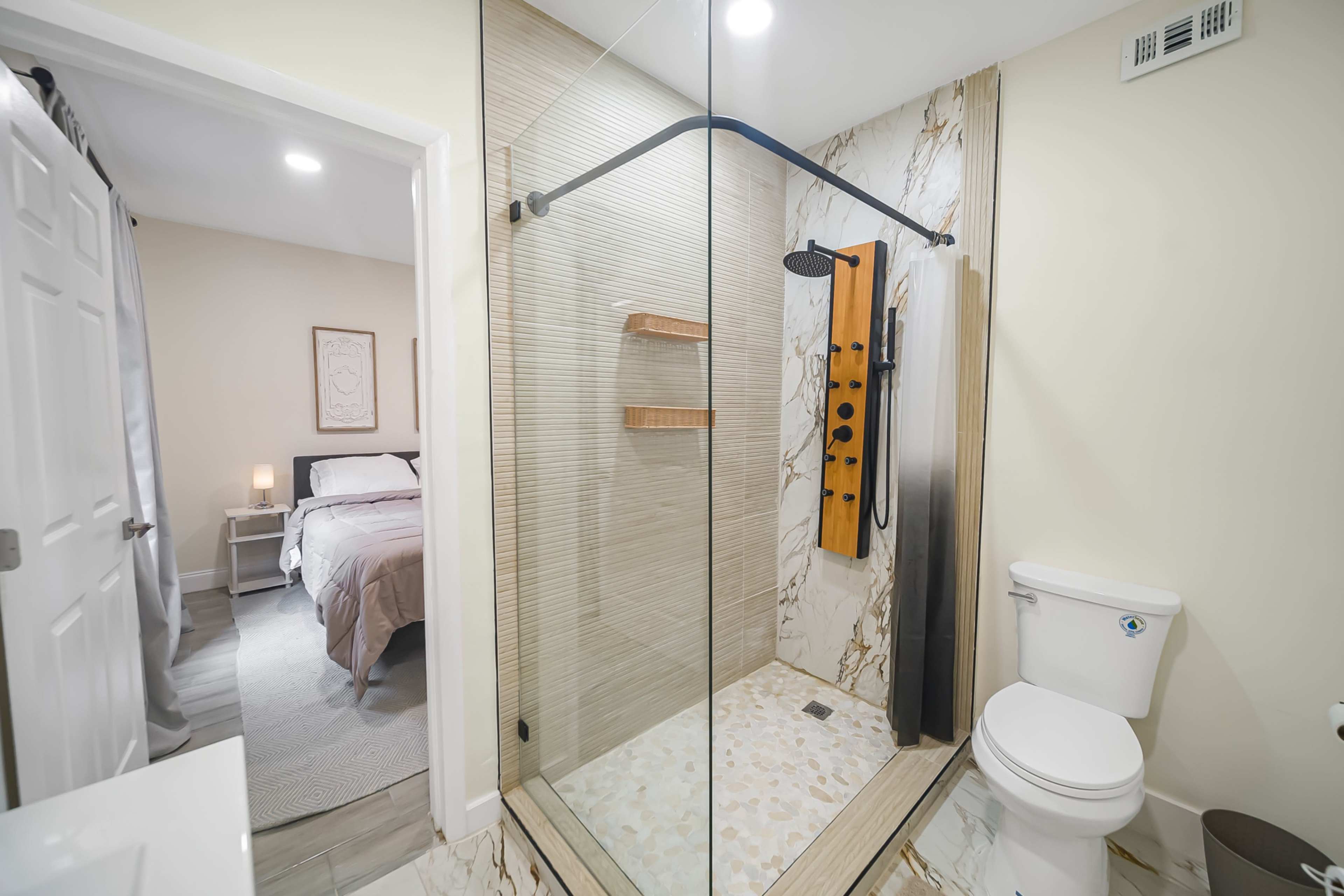 The image shows a modern bathroom featuring a glass-enclosed shower, a toilet, and views of a bedroom through an open doorway.