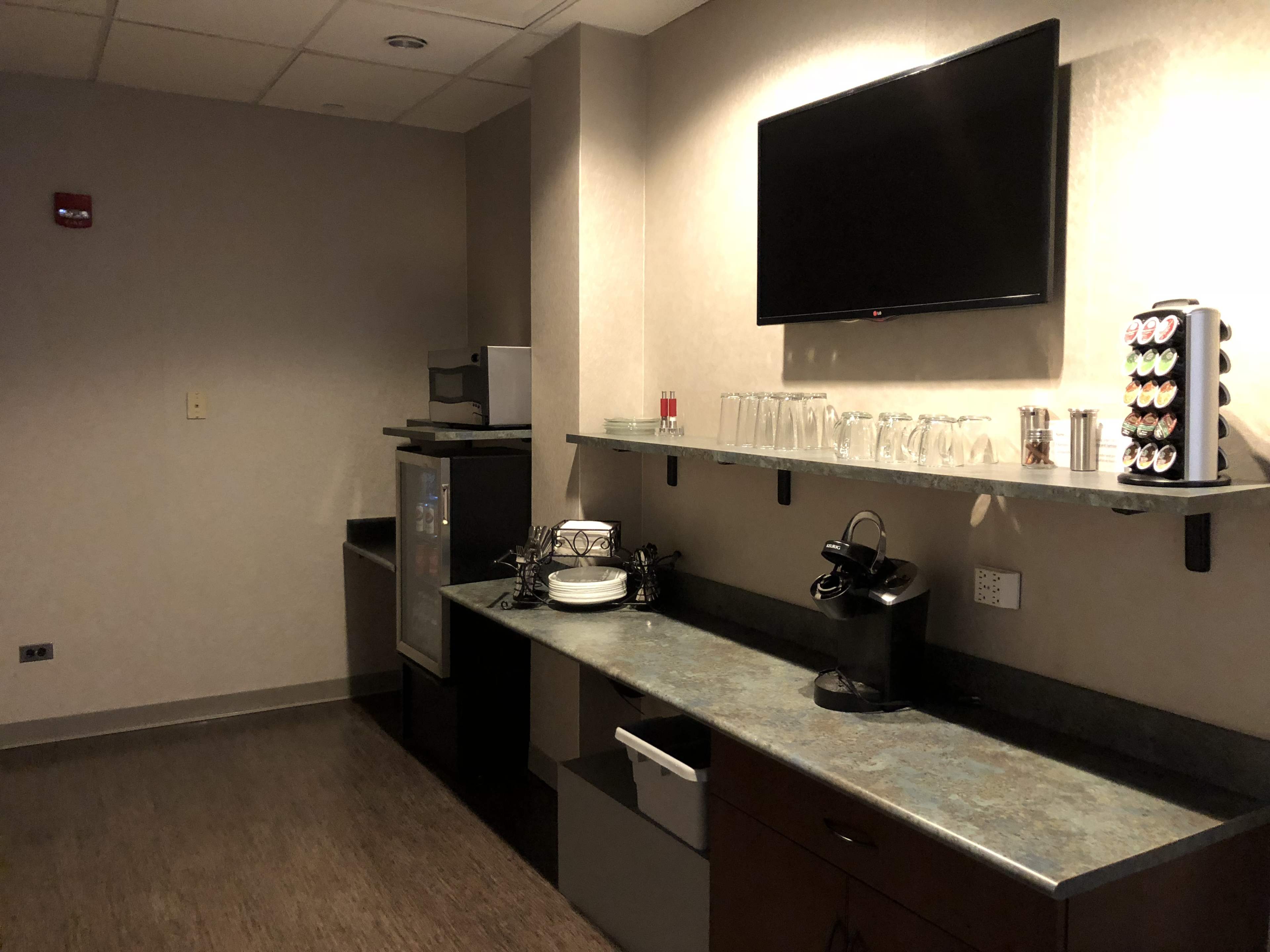 The image shows a small kitchenette with a countertop, sink, a coffee maker, glasses, and a refrigerator against a plain wall.