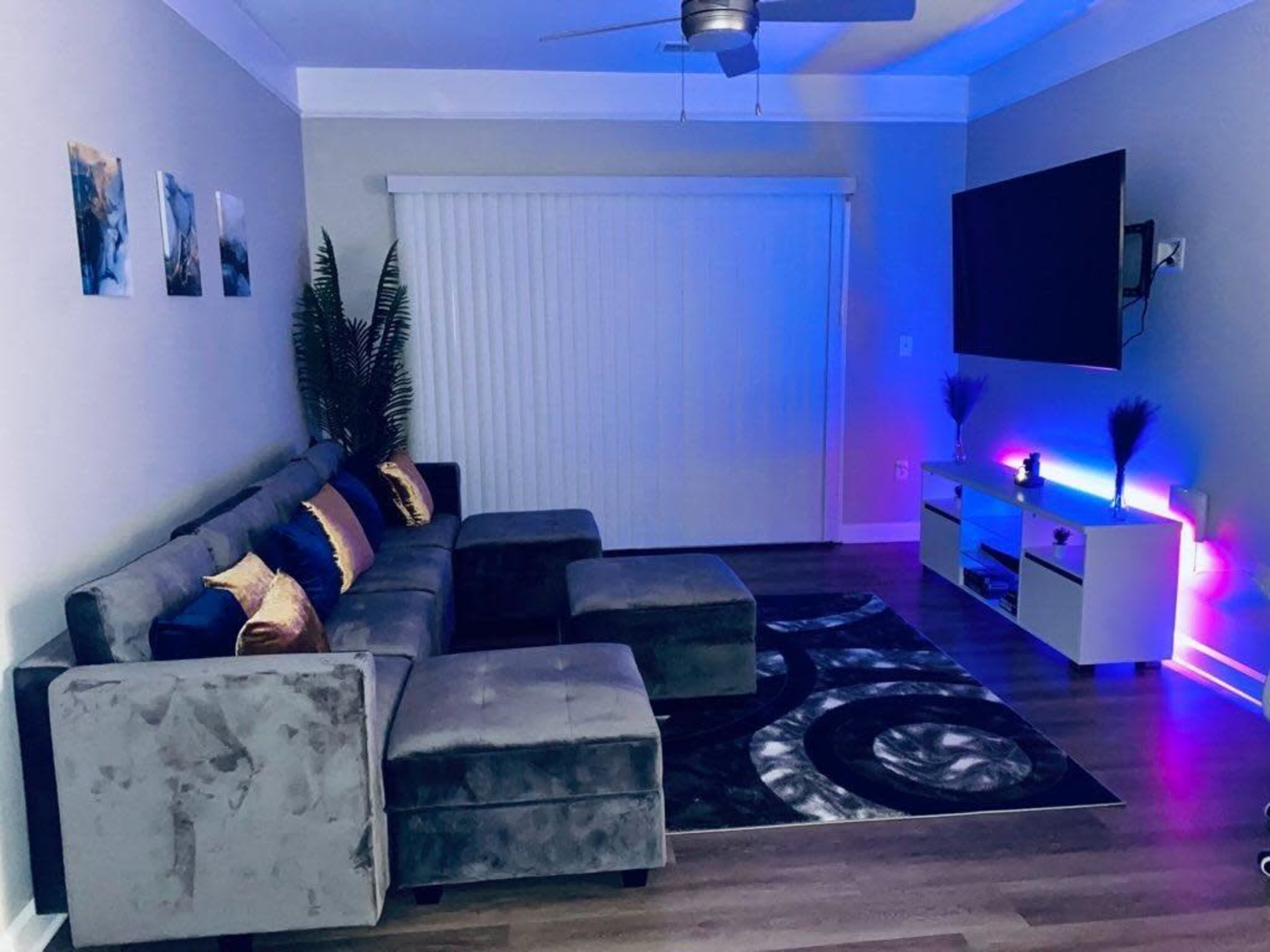 A modern living room features a gray sectional sofa, a white entertainment unit with colored lighting, and a large television against a neutral wall.