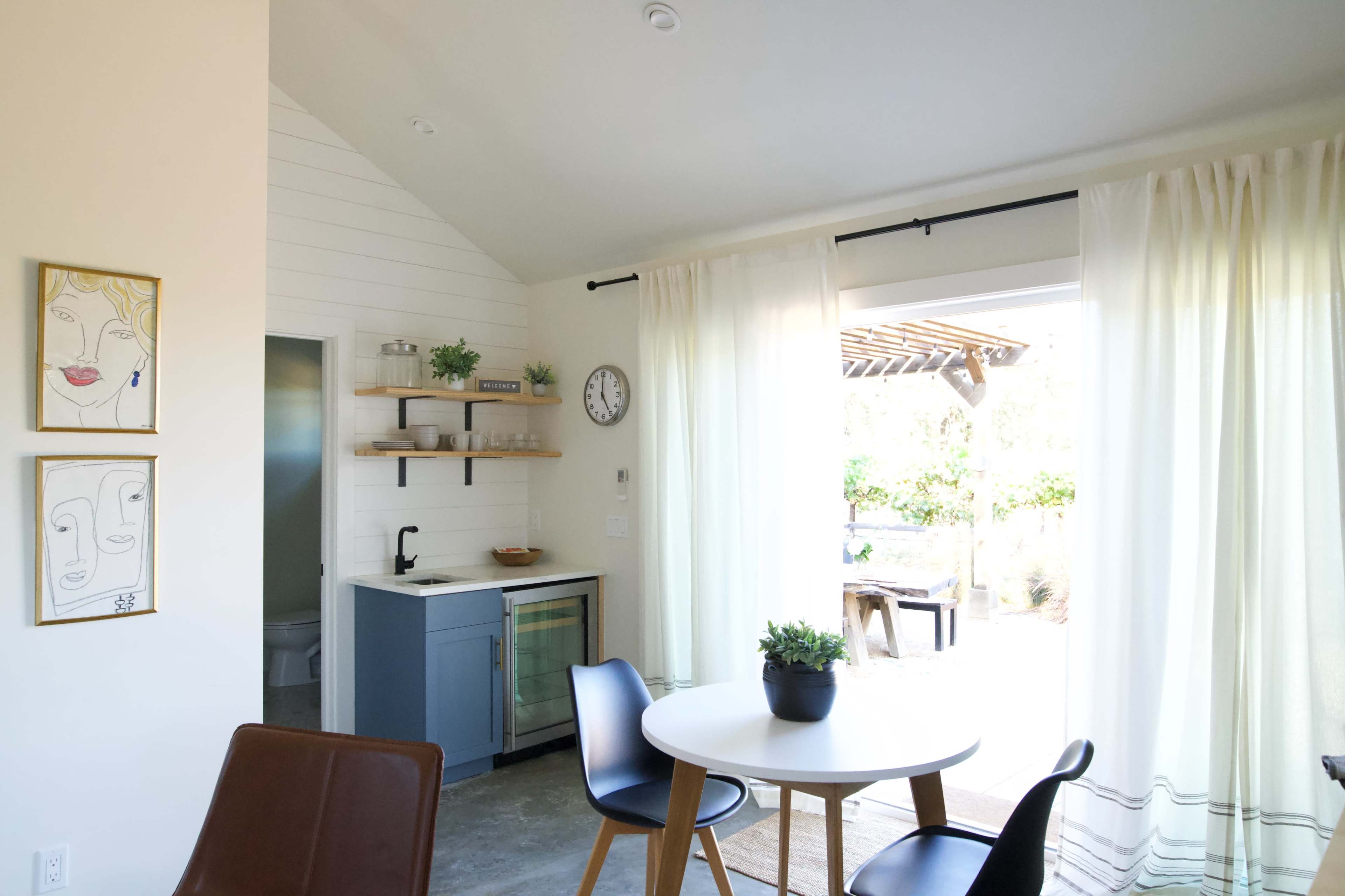 A modern kitchen area features a round table with chairs, open shelving, and large windows that lead to an outdoor space.