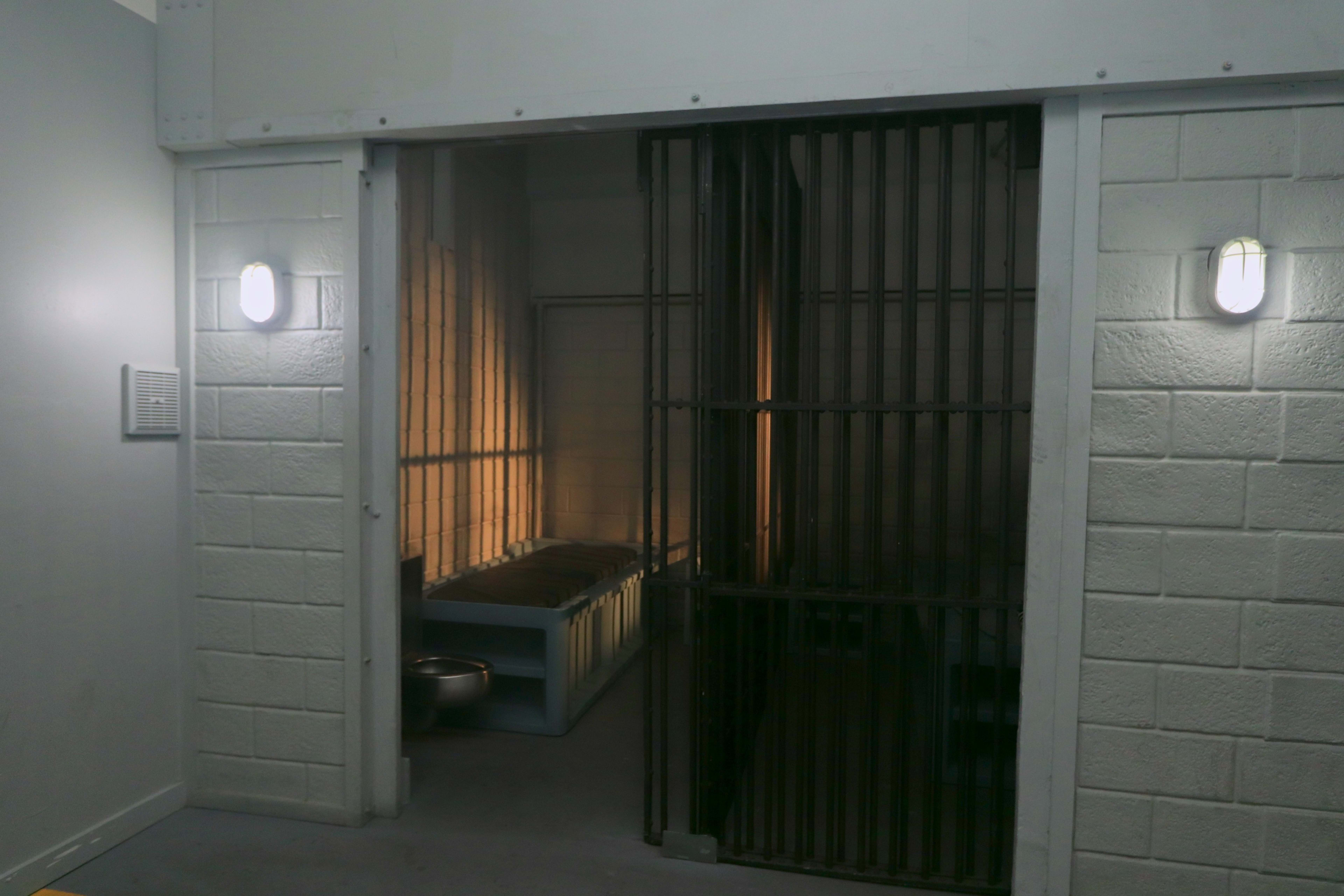 The image shows the interior of a prison cell, featuring a barred gate, a bed, and a small metal toilet.