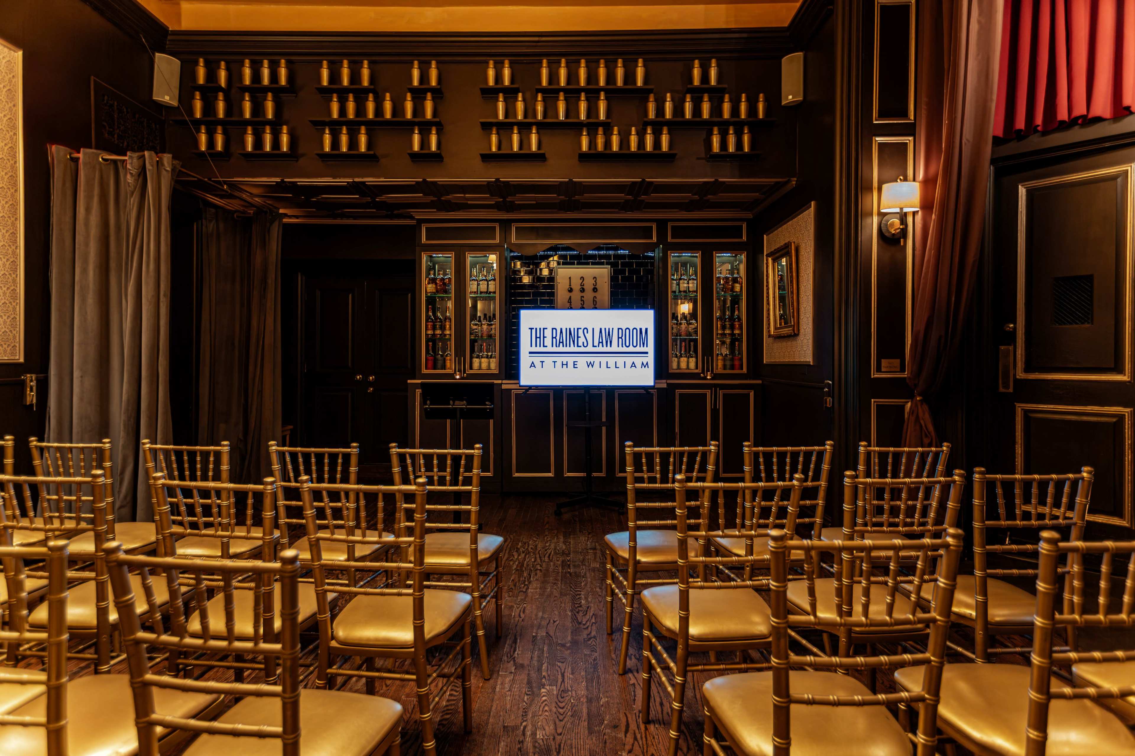 The image shows a dimly lit room with rows of gold chairs facing a large illuminated sign that reads "THE JAMES LAV ROOM AT THE WILLIAM," against a backdrop of wooden shelves filled with bottles.