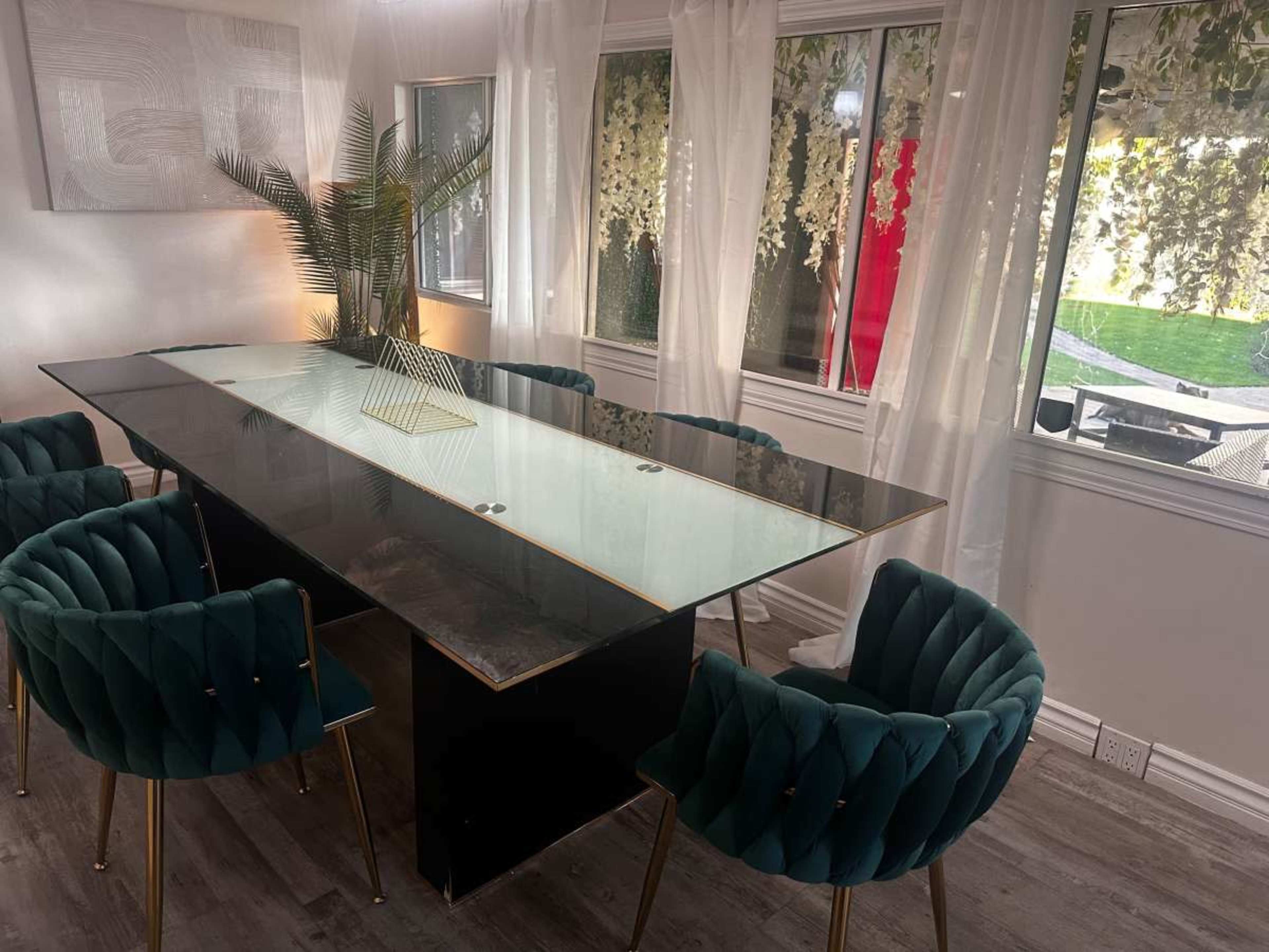 A modern dining area features a sleek glass-top table surrounded by green velvet chairs and large windows letting in natural light.